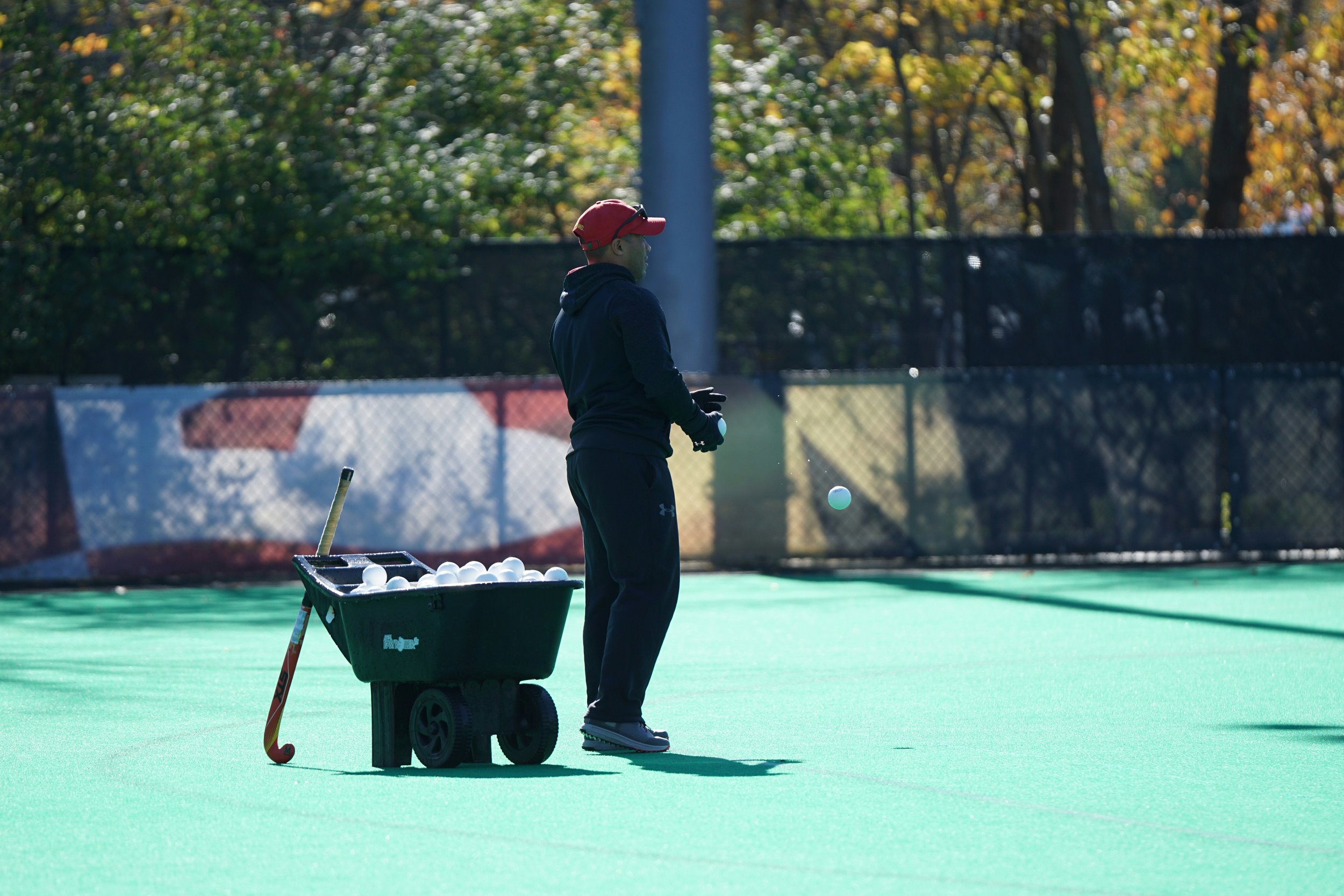 Person wearing a red cap and black hoodie practicing golf on a green outdoor course with a cart full of white golf balls nearby.