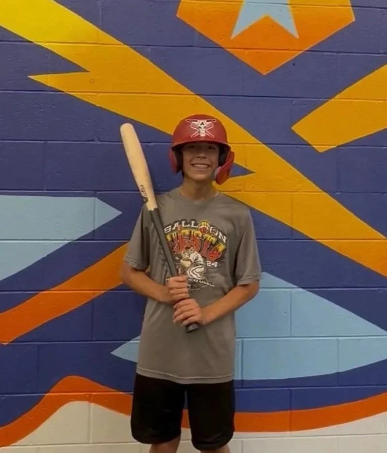 A young boy wearing a red baseball helmet with a sticker of a cow skull, holding a baseball bat, smiling, standing in front of a brightly colored mural with yellow, blue, orange, and light blue geometric shapes on a wall.
