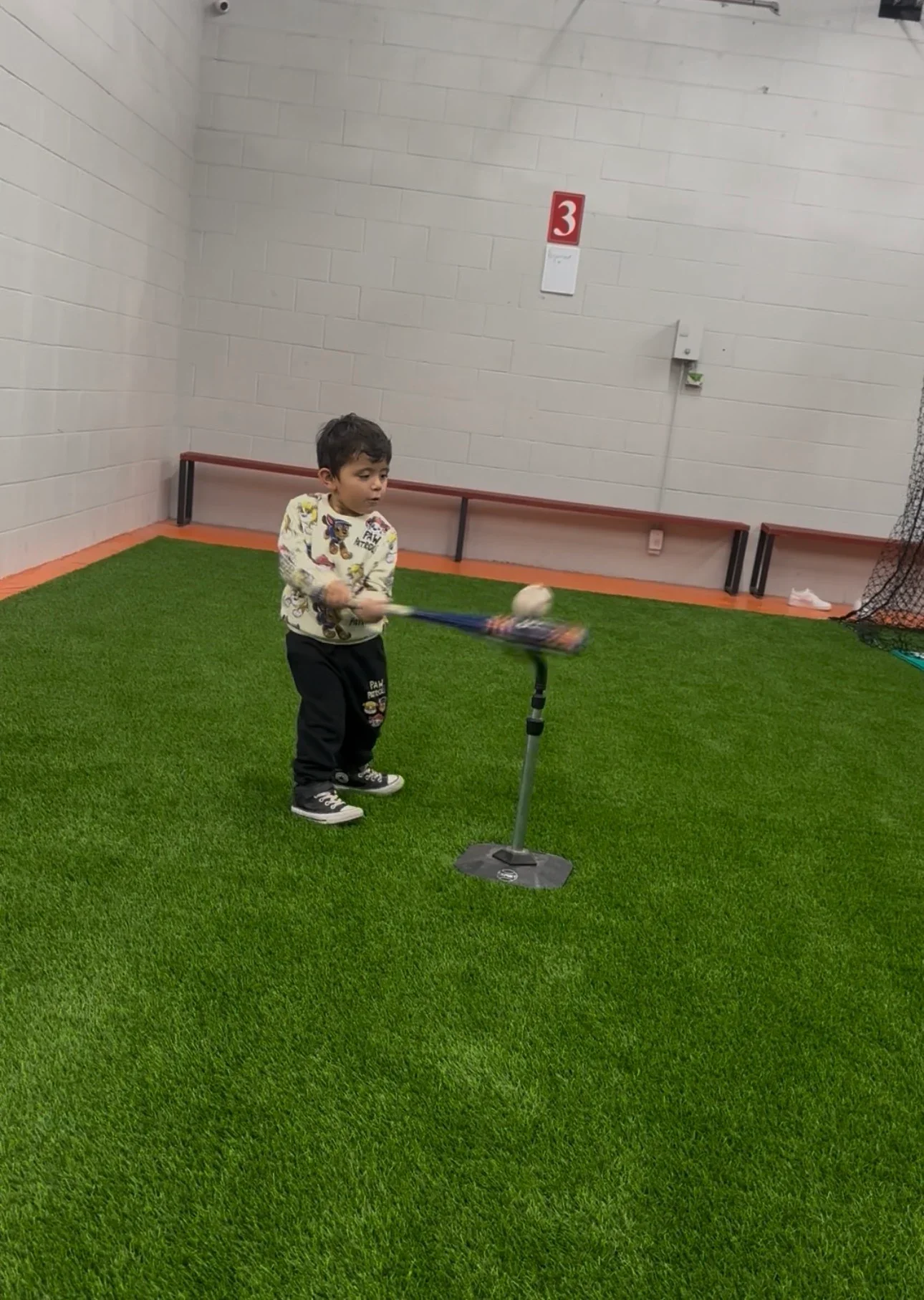 A young boy practicing baseball indoors on artificial green turf, swinging a bat at a ball on a tee.