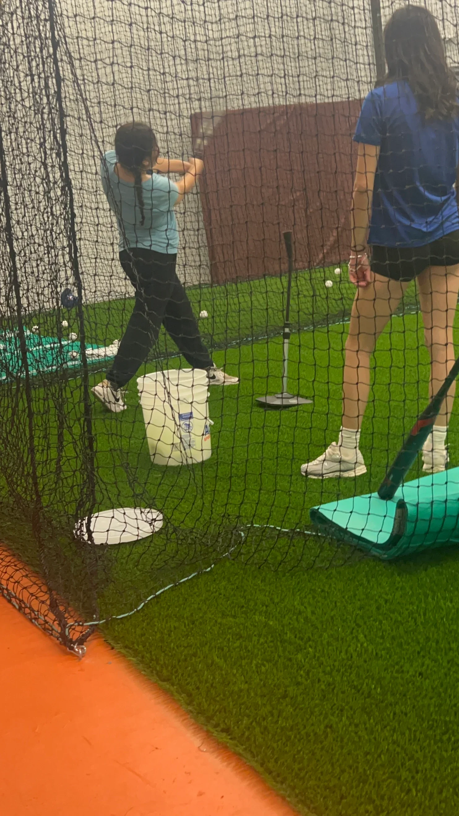 Two women practicing batting skills in an indoor batting cage, with one woman swinging a bat and the other watching, surrounded by equipment like balls, a tee, and batting mats.