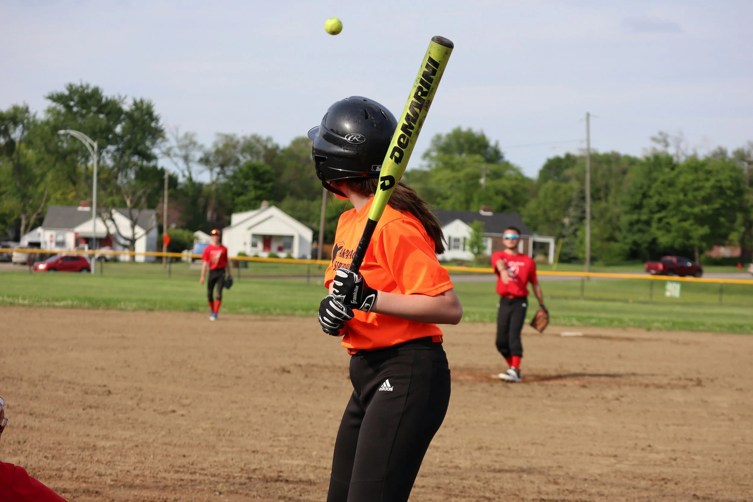 A young softball player in an orange shirt and black helmet standing at home plate, holding a yellow Bat Recurve, as a ball is pitched toward her. Infield and outfield players are visible in the background on the grassy field with houses and trees beyond.