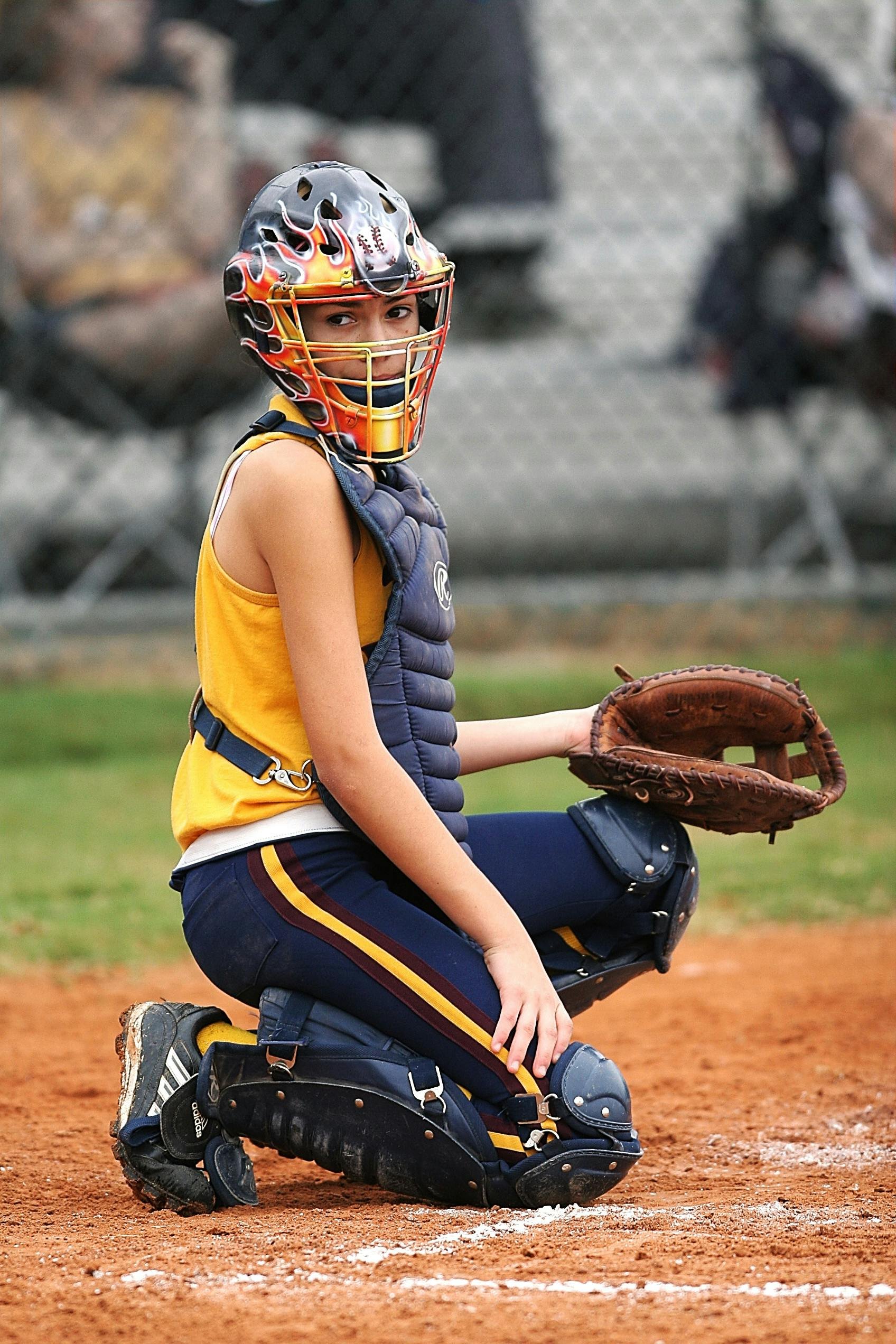 A female youth baseball or softball catcher in full gear, squatting on the field with a glove, wearing a helmet with a face cage, chest protector, and padded pants, on a dirt field.