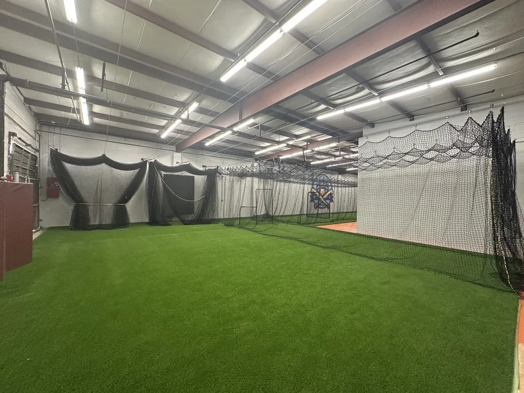 Indoor baseball practice area with artificial turf, nets, and pitching lanes.