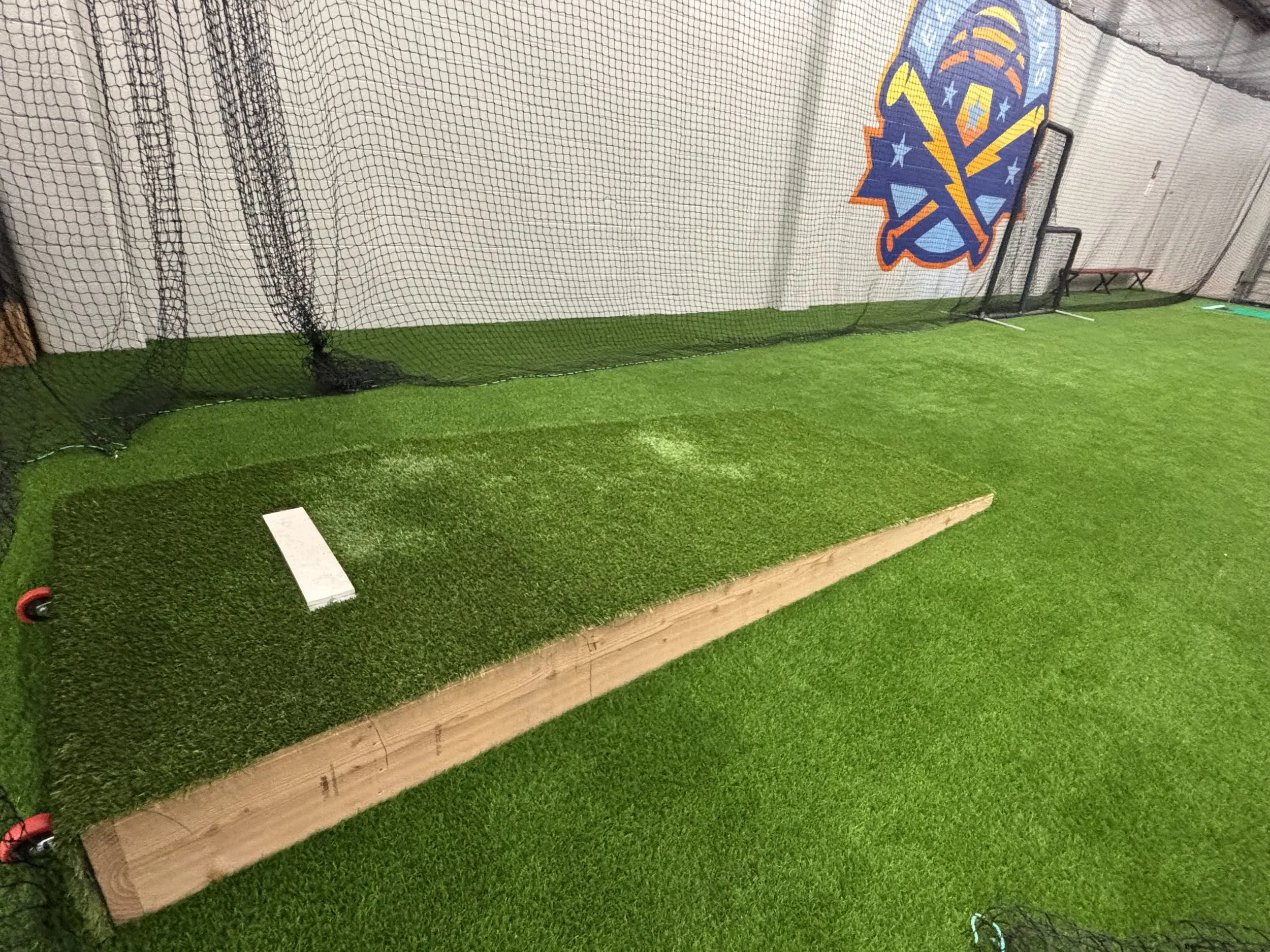 Indoor sports facility with artificial green turf, a batting tee, and a backstop with a sports logo on the wall.