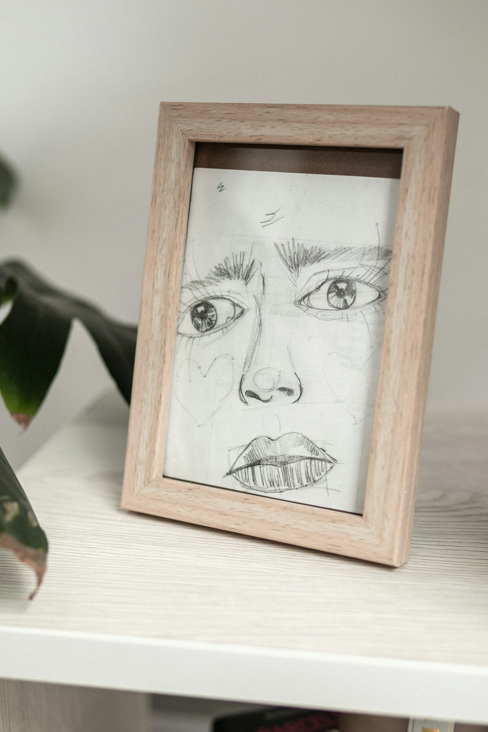 A sketch of a woman's face with detailed eyes, nose, and lips inside a wooden frame. A plant is partially visible on the left.