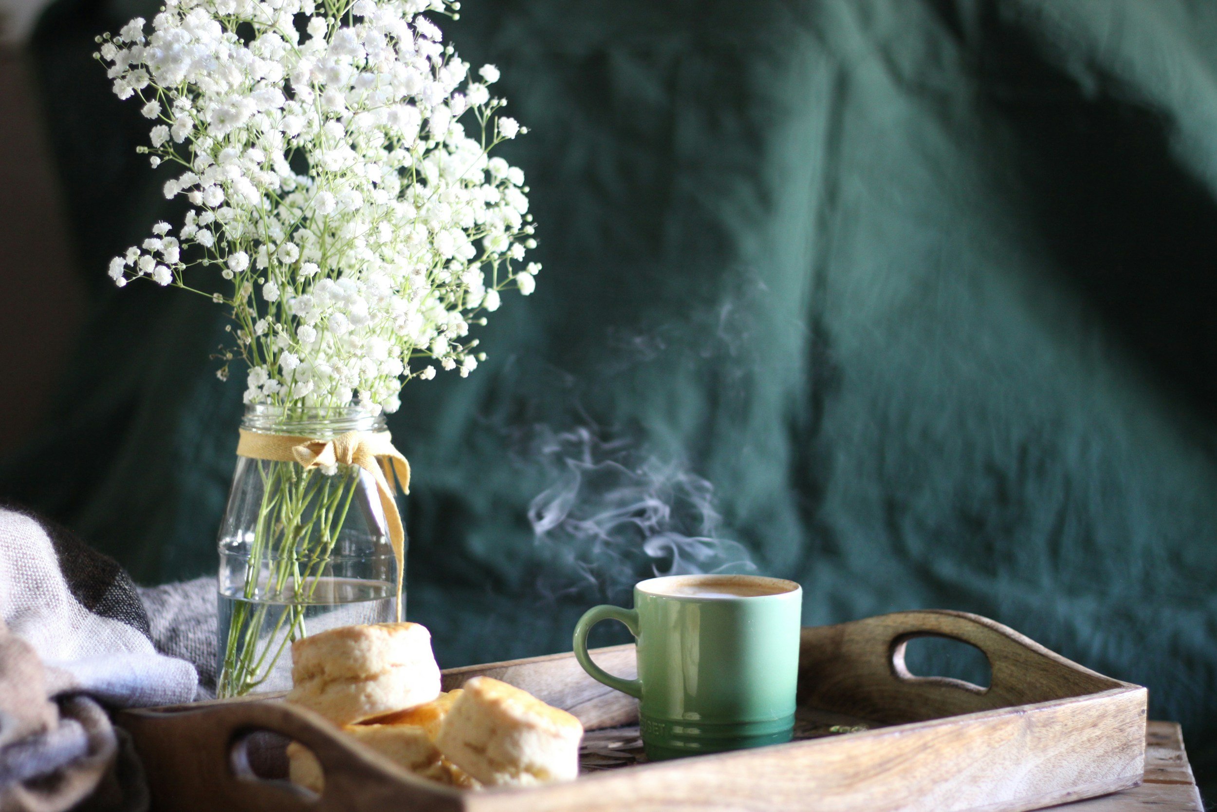 A still life of a wooden tray with a steaming cup of coffee, a jar of white flowers, and some pastries on a table, with a dark green background.