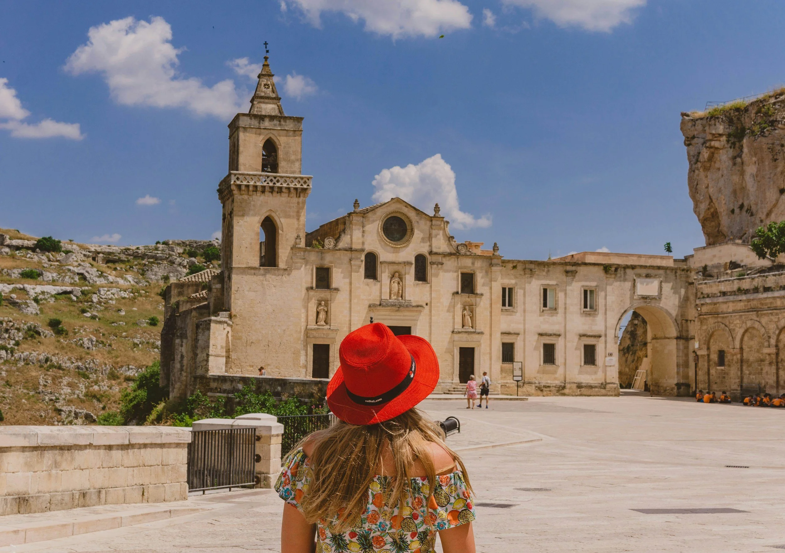 A young woman with long hair, wearing a red hat and a colorful off-the-shoulder dress, stands in front of an old stone church with a bell tower, under a partly cloudy sky.