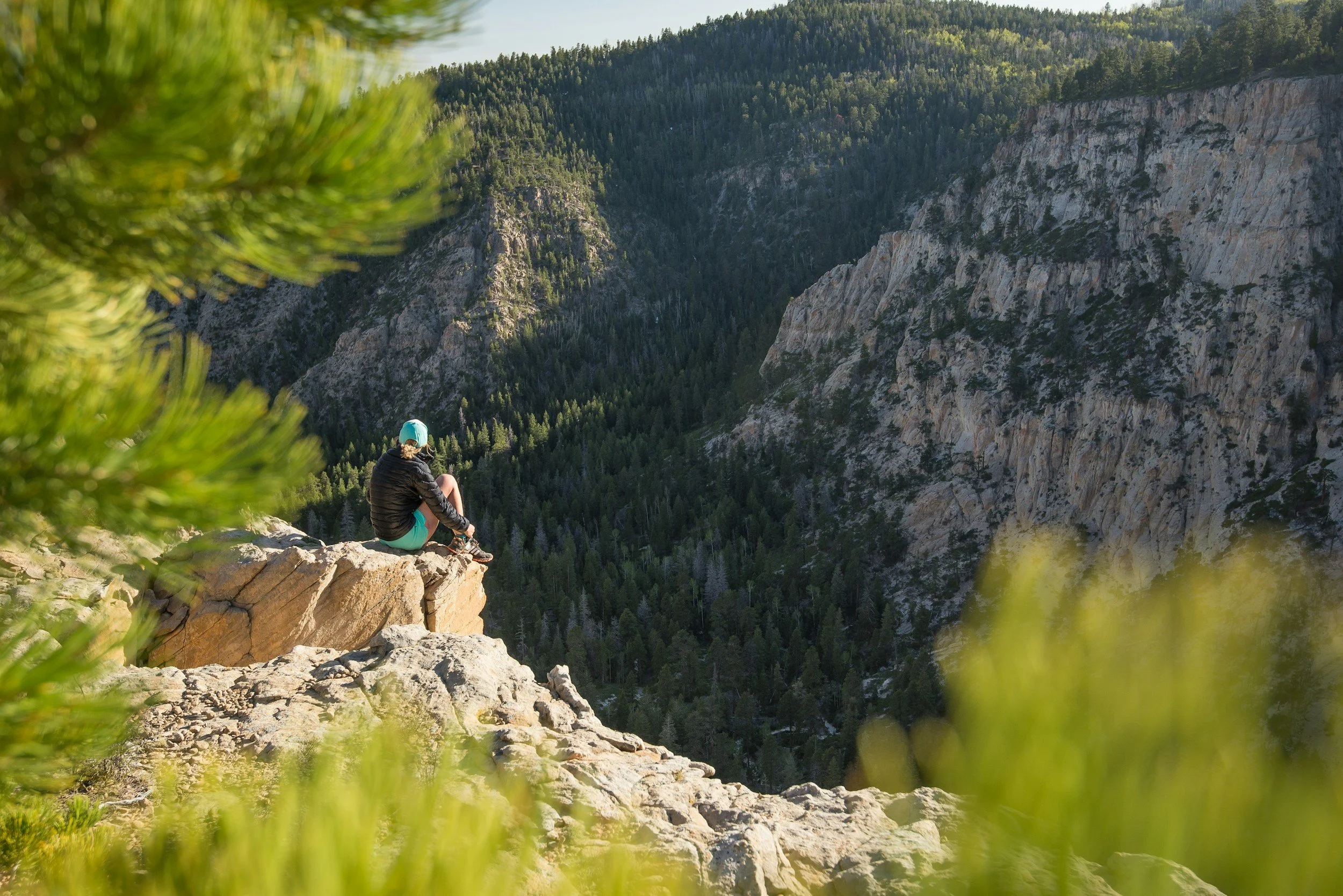 A person sitting on a rocky ledge overlooking a valley with forested mountains in the distance.