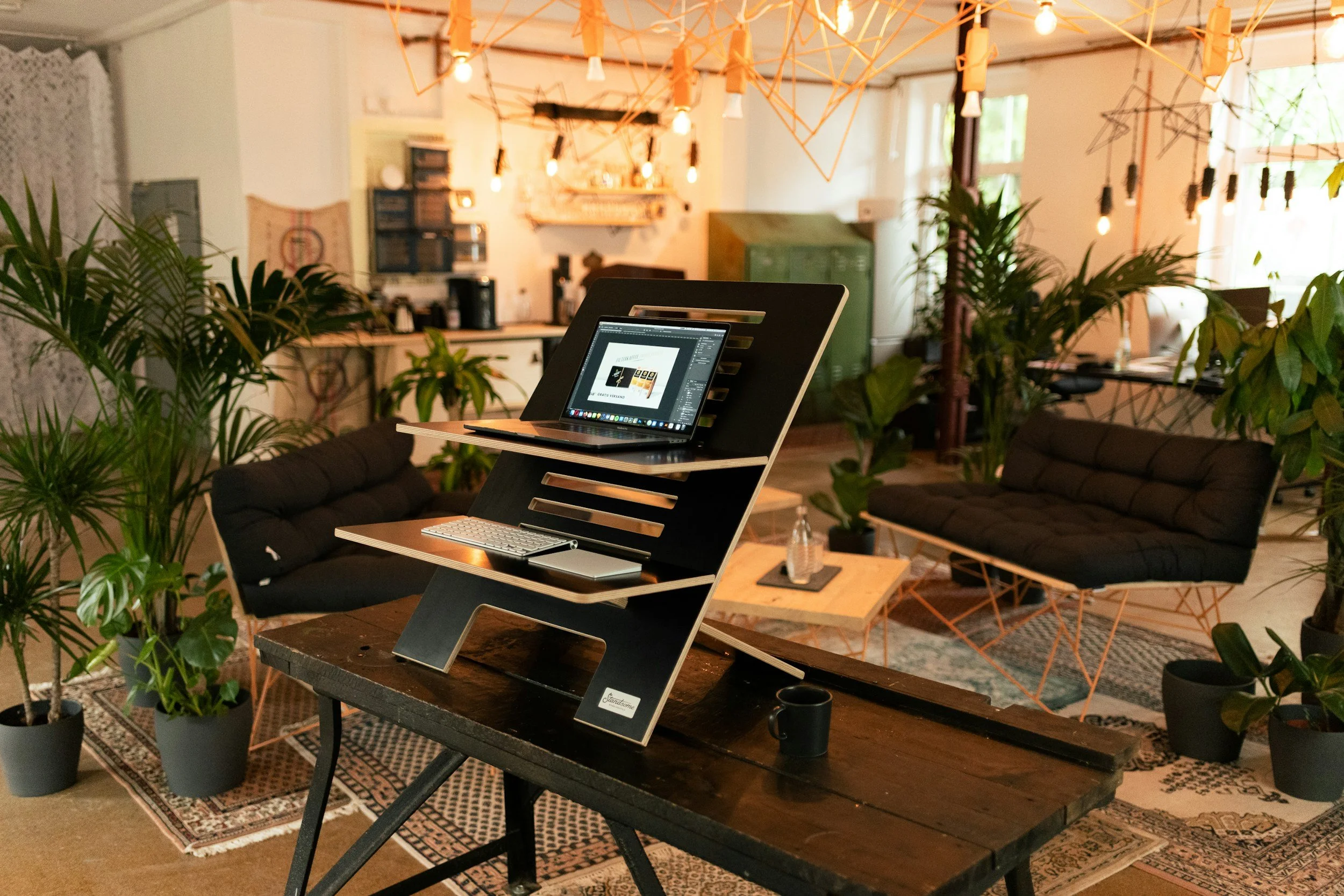 Interior of a cozy, modern office or lounge space with plants, black cushioned sofas, wooden tables, and warm lighting hanging from the ceiling.