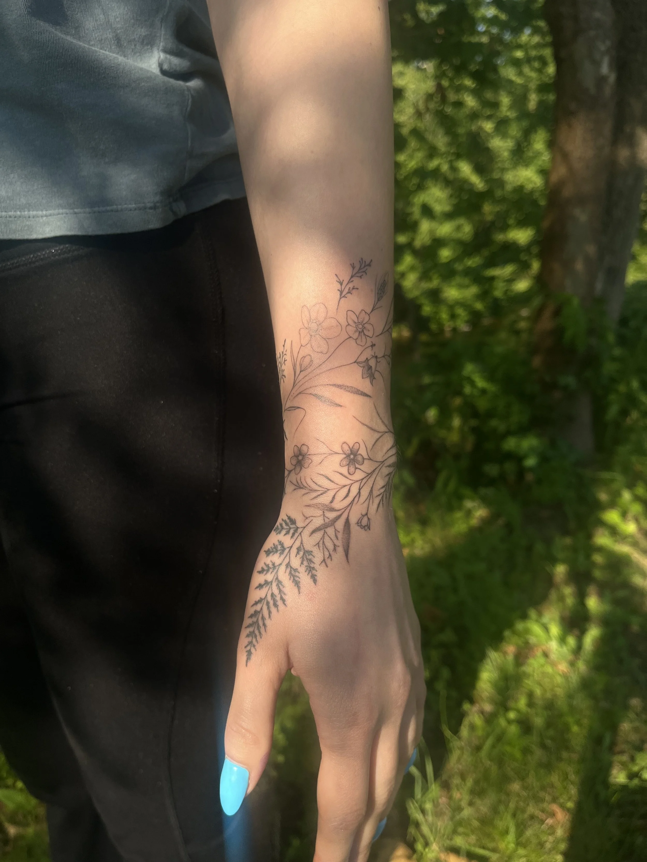 A person's arm and hand with a fine line floral tattoo design, outdoors with green foliage in the background, wearing a gray shirt and black pants.