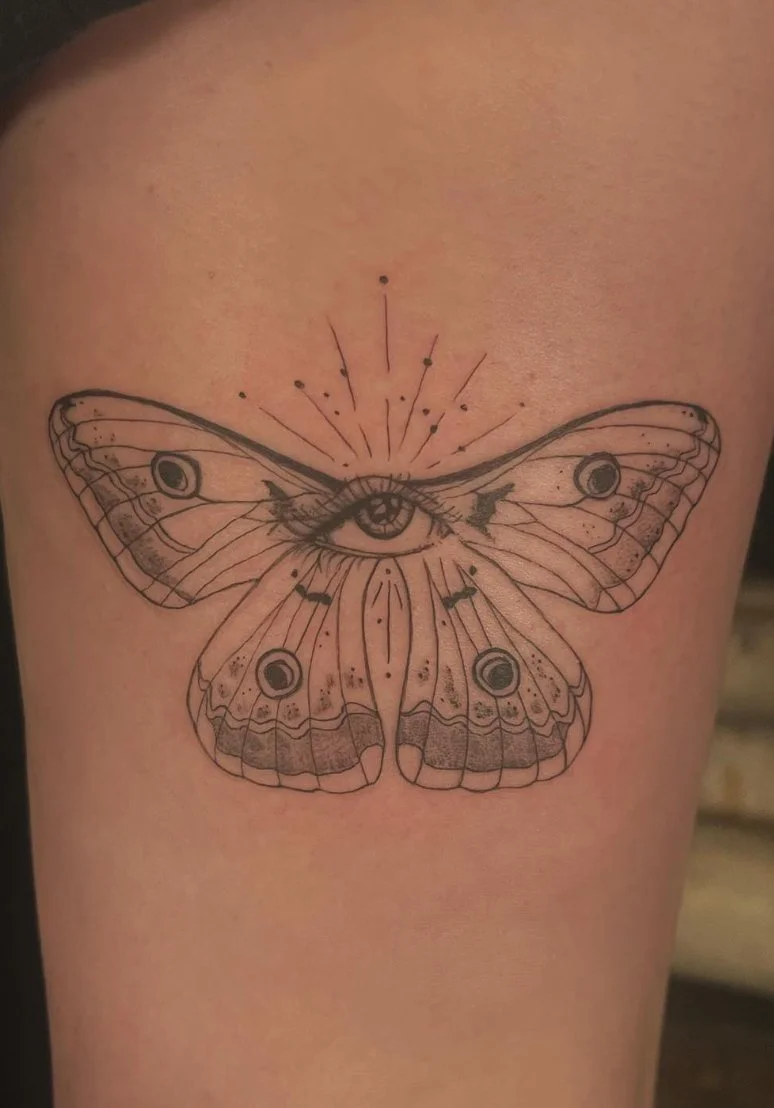 Tattoo of a butterfly with an eye in the center of its body and celestial designs, including stars and rays, on its wings.