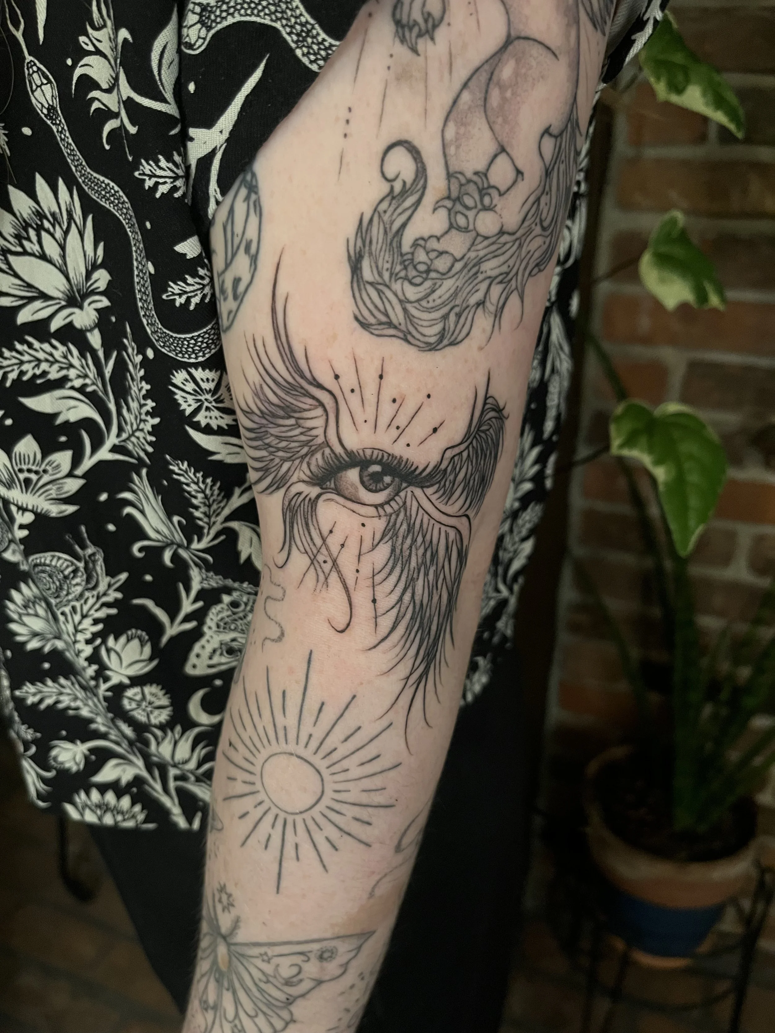 Tattoo sleeve featuring an eye with detailed eyelashes and rays emanating from it, a sun symbol, and various celestial and mystical designs on a person's arm.