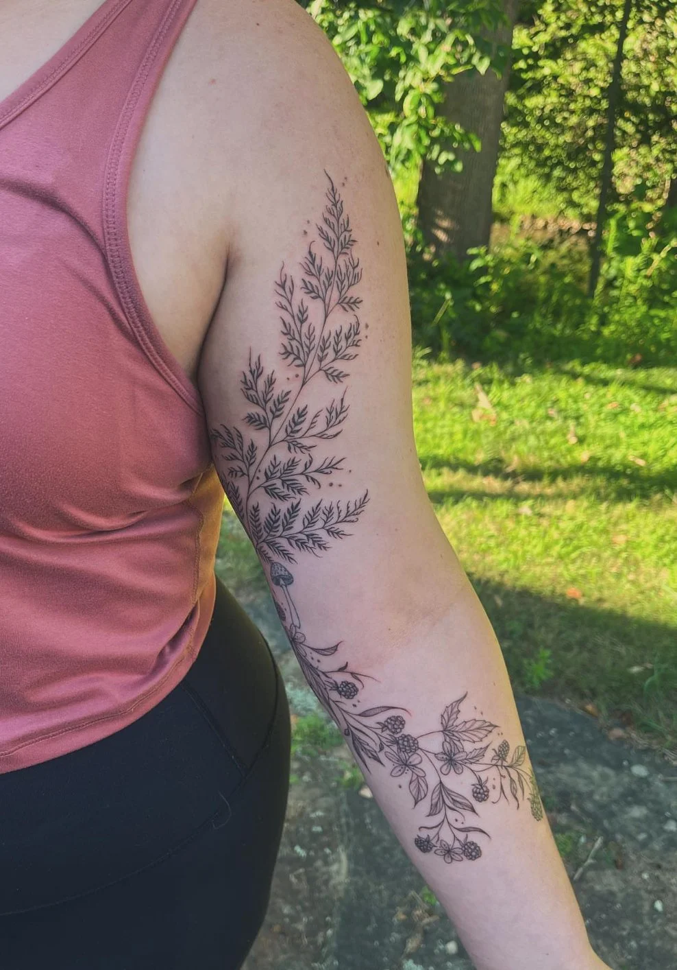A person with a large black ink botanical tattoo on their left upper arm. The tattoo features detailed fern-like leaves and flowers. The person is wearing a sleeveless pink shirt and black pants, outdoors with green trees and grass in the background.