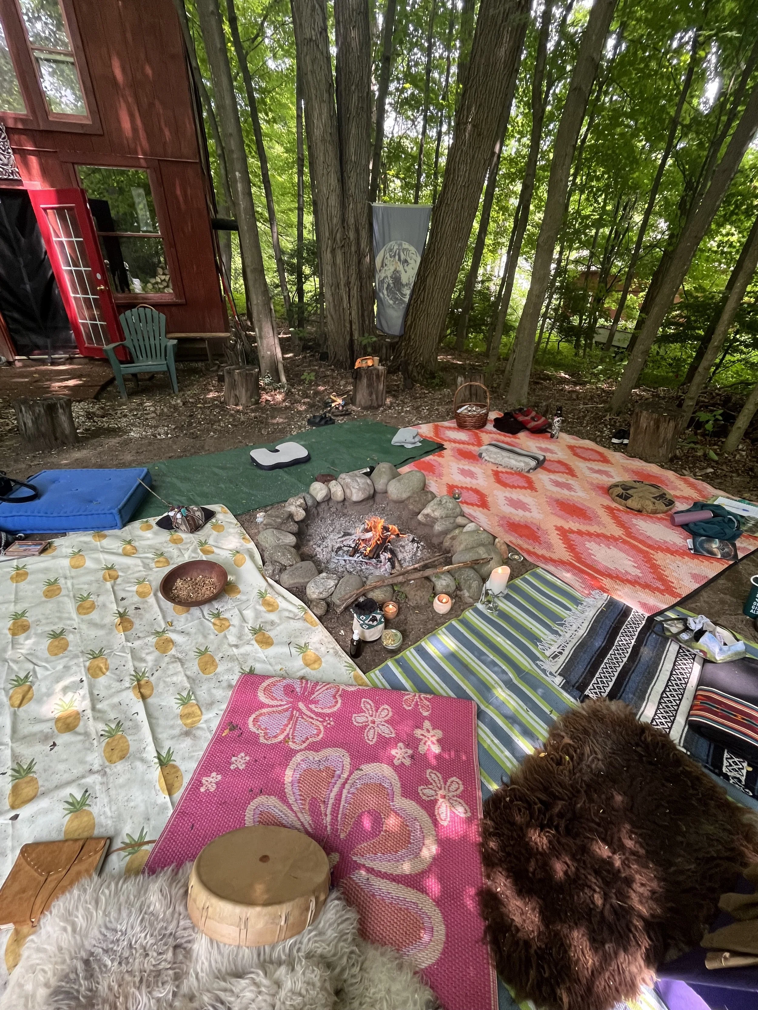 A wooded outdoor camping scene with a small campfire surrounded by various colorful blankets, mats, and pillows on the ground. This is a women's circle space, a sacred temple where women gather.