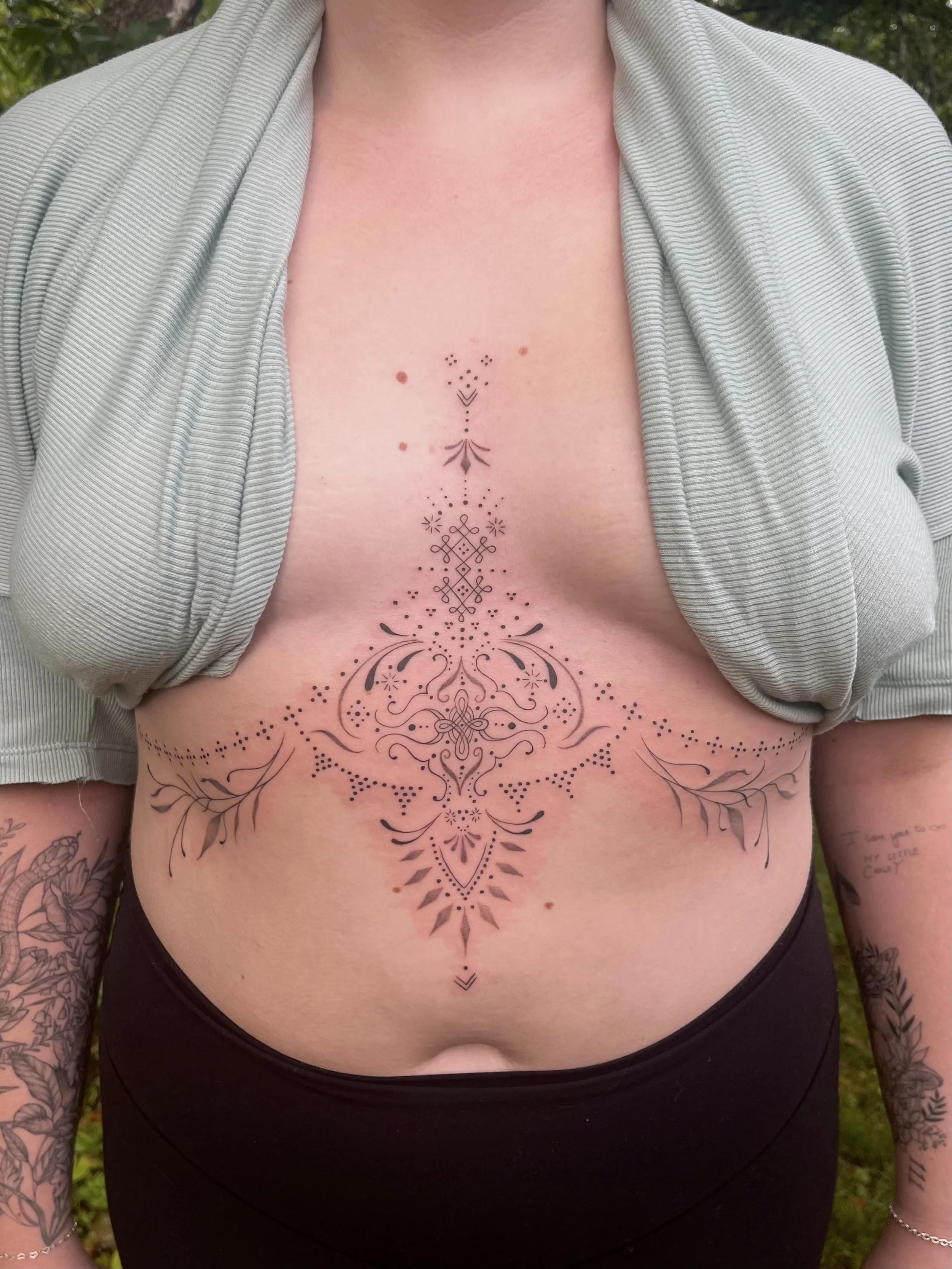 A person with tattoos on their chest and arms, wearing a light gray open shirt, showing a symmetrical black tattoo with intricate geometric and floral patterns on their upper chest and torso.