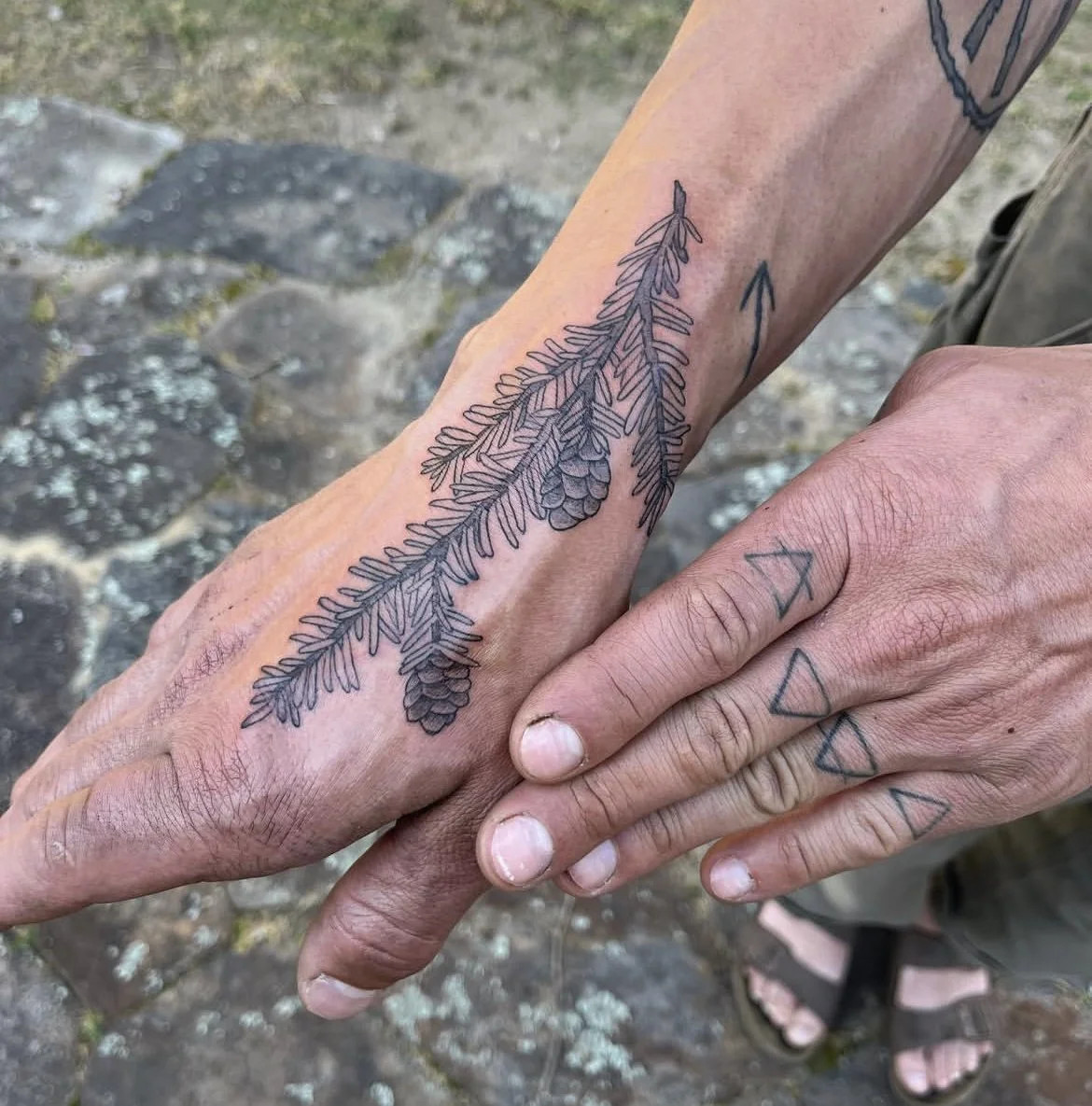 Close-up of two hands, one with a detailed tattoo of pine branches and pinecones and the other with simple geometric sacred elements tattoos