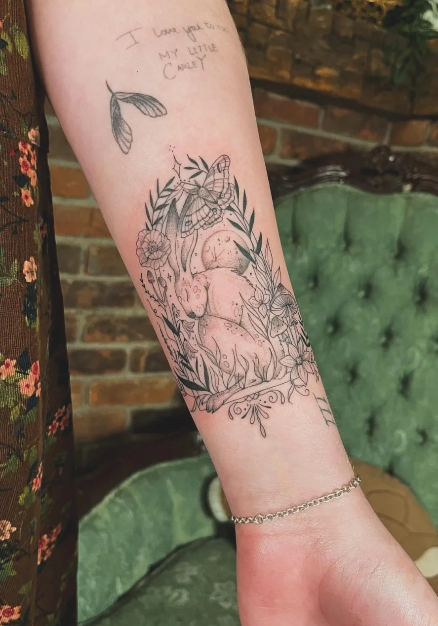A tattoo on a person's forearm featuring a moon, surrounded by flowers, leaves, a butterfly, and a moth. The tattoo includes a line drawing of a small creature with a smiling face. Above the tattoo, there are handwritten words that read, 'I love you to the moon and my little carey.'