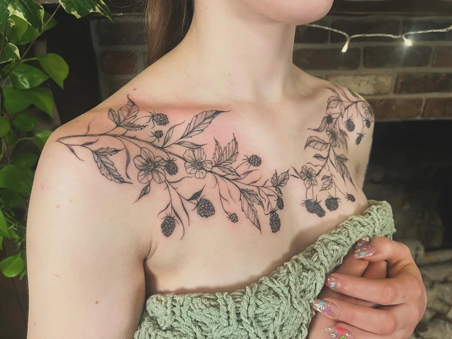 A woman with a botanical vine floral tattoo on her chest featuring blackberry branches, flowers, and leaves. She is holding a green textured towel near her chest.