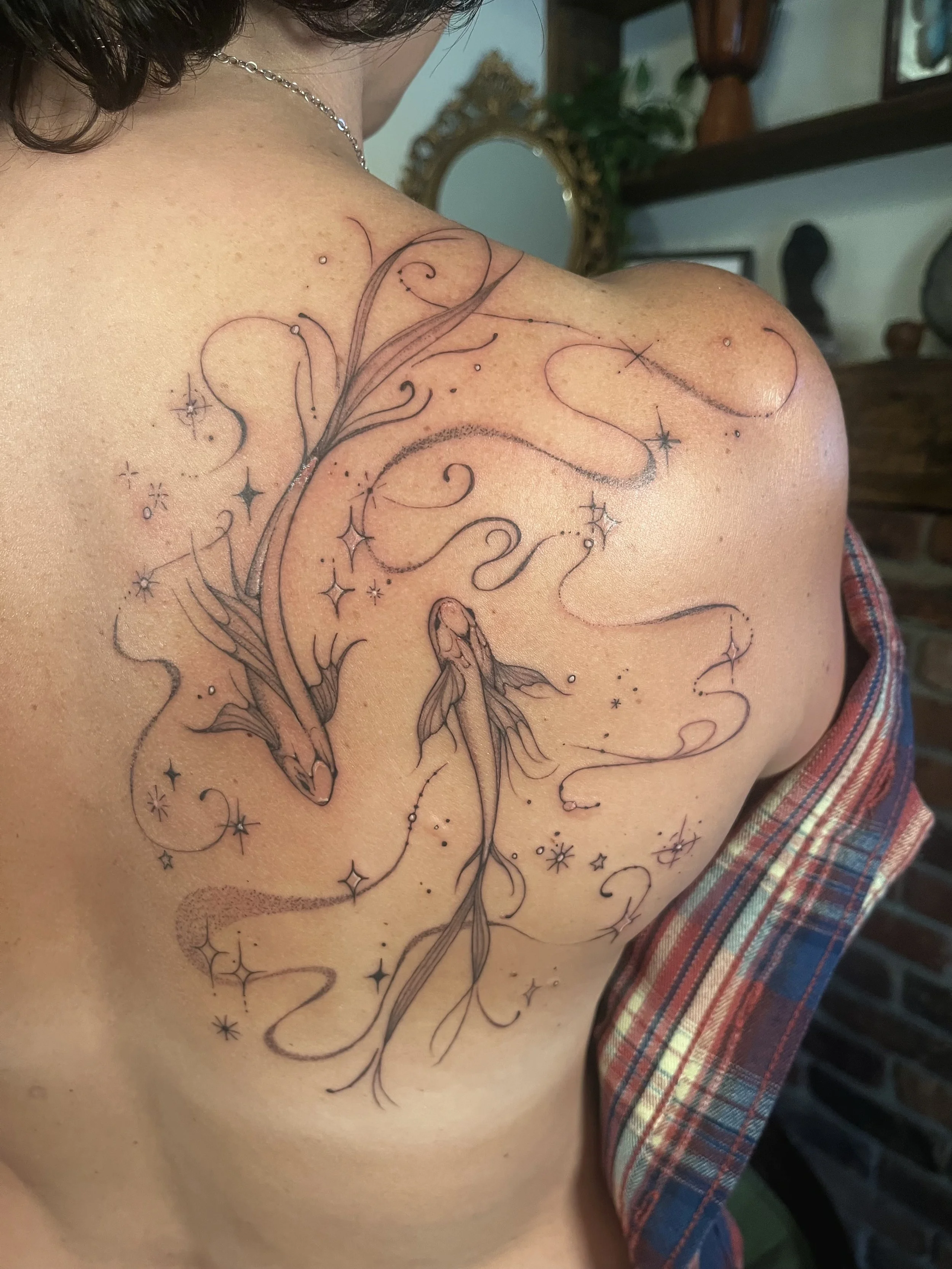 An astrology tattoo of two Pisces fish swimming around each other, surrounded by whimsical stars and swirling lines on a person's shoulder.