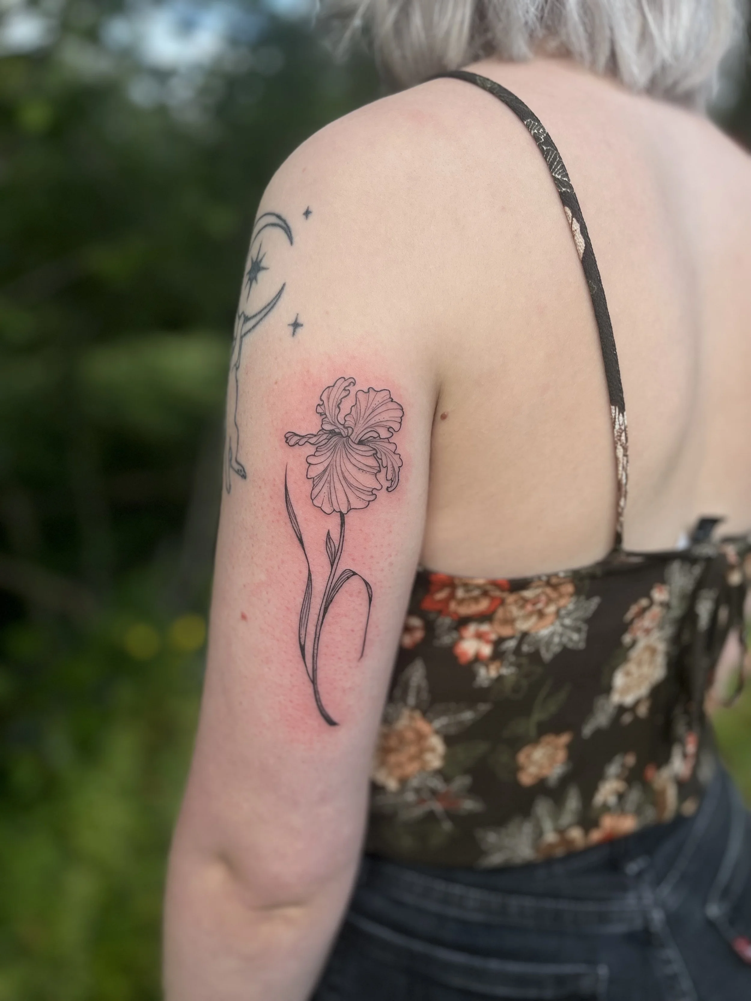 A woman with short blonde hair showing a tattoo of a flower on her upper arm, with some moon and star tattoos nearby, outdoors with greenery in the background.