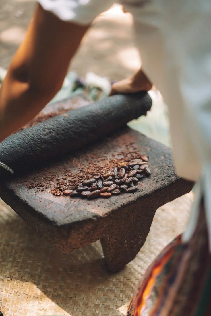 A person is roasting coffee beans on an outdoor stone grinder, with coffee beans scattered on it, near a body of water.