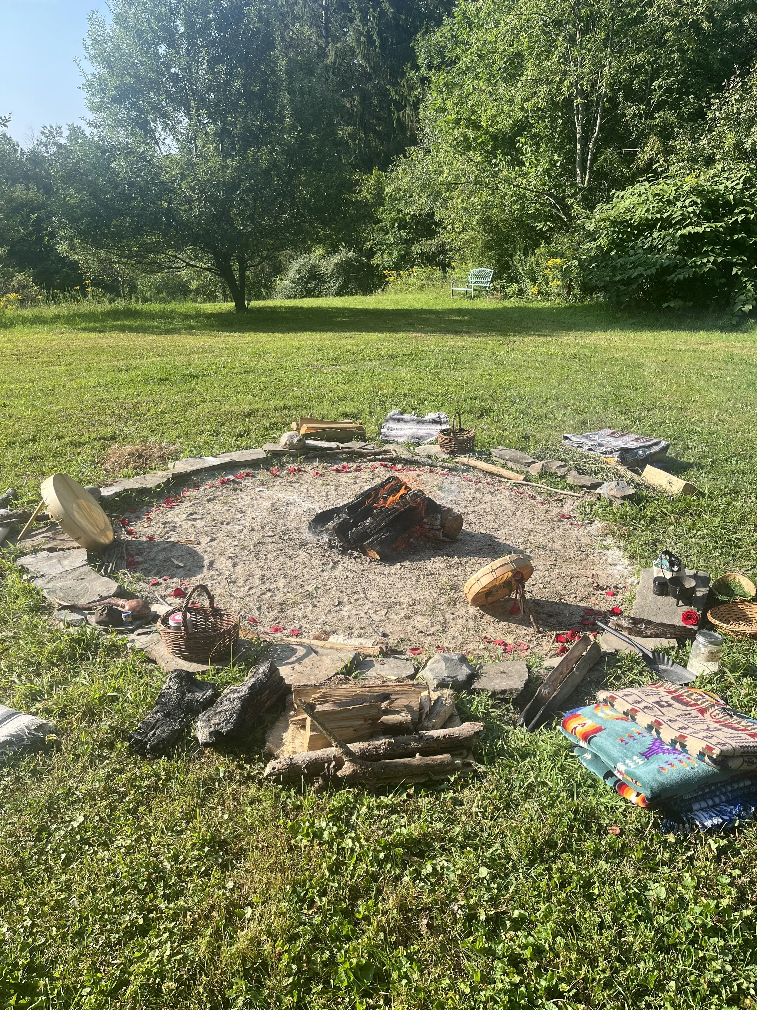 Outdoor scene of a fire pit with scattered towels, baskets, and firewood, surrounded by grass and trees in a park or backyard setting. This is a sacred ritual where drum circle, sacred song and healing take place.