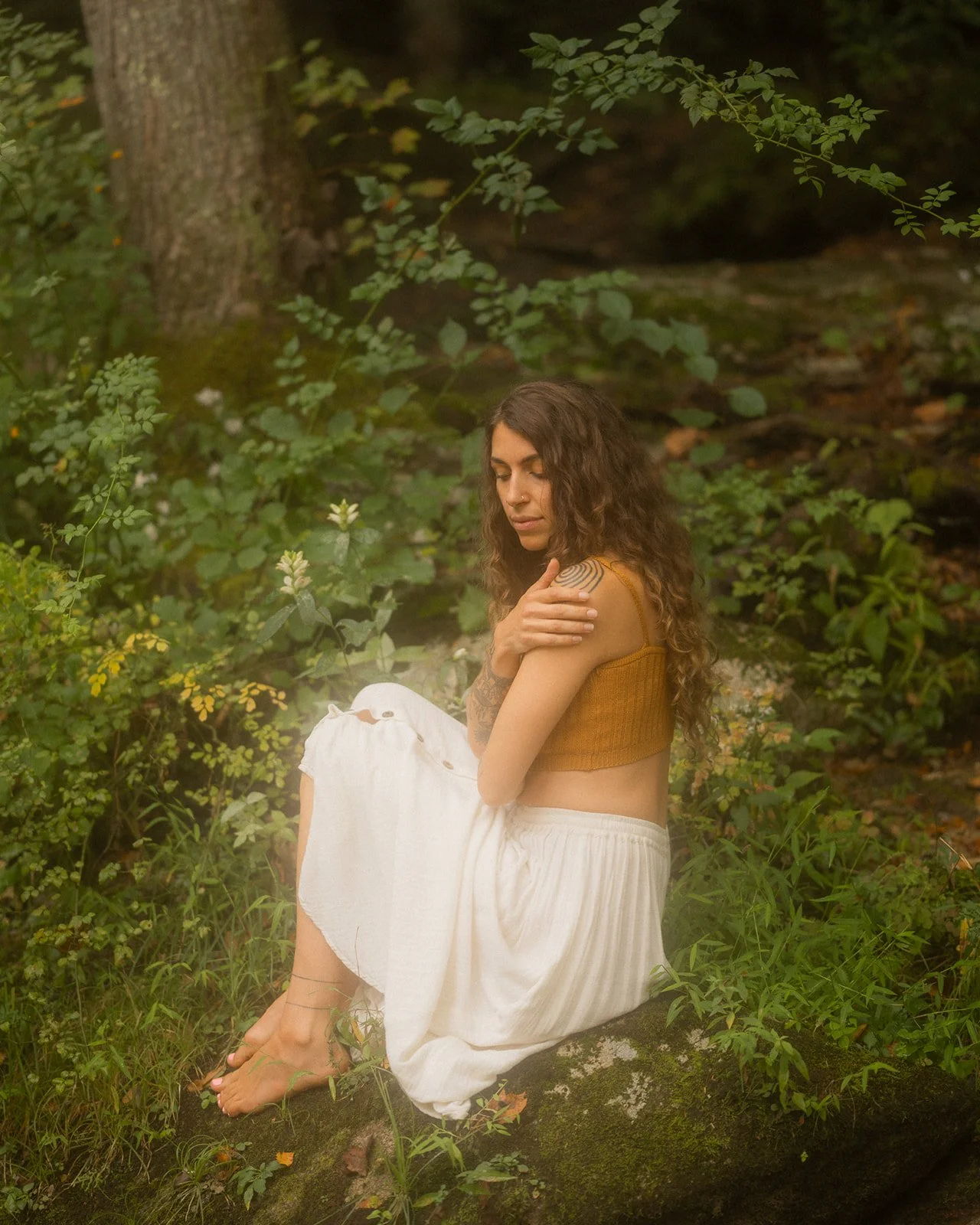 A woman with long curly hair, wearing a mustard yellow top and white pants, sits on a moss-covered rock in a lush green forest, with her eyes closed and arms crossed over her shoulders. She is a tattoo artist and owner of root and ritual tattoo.