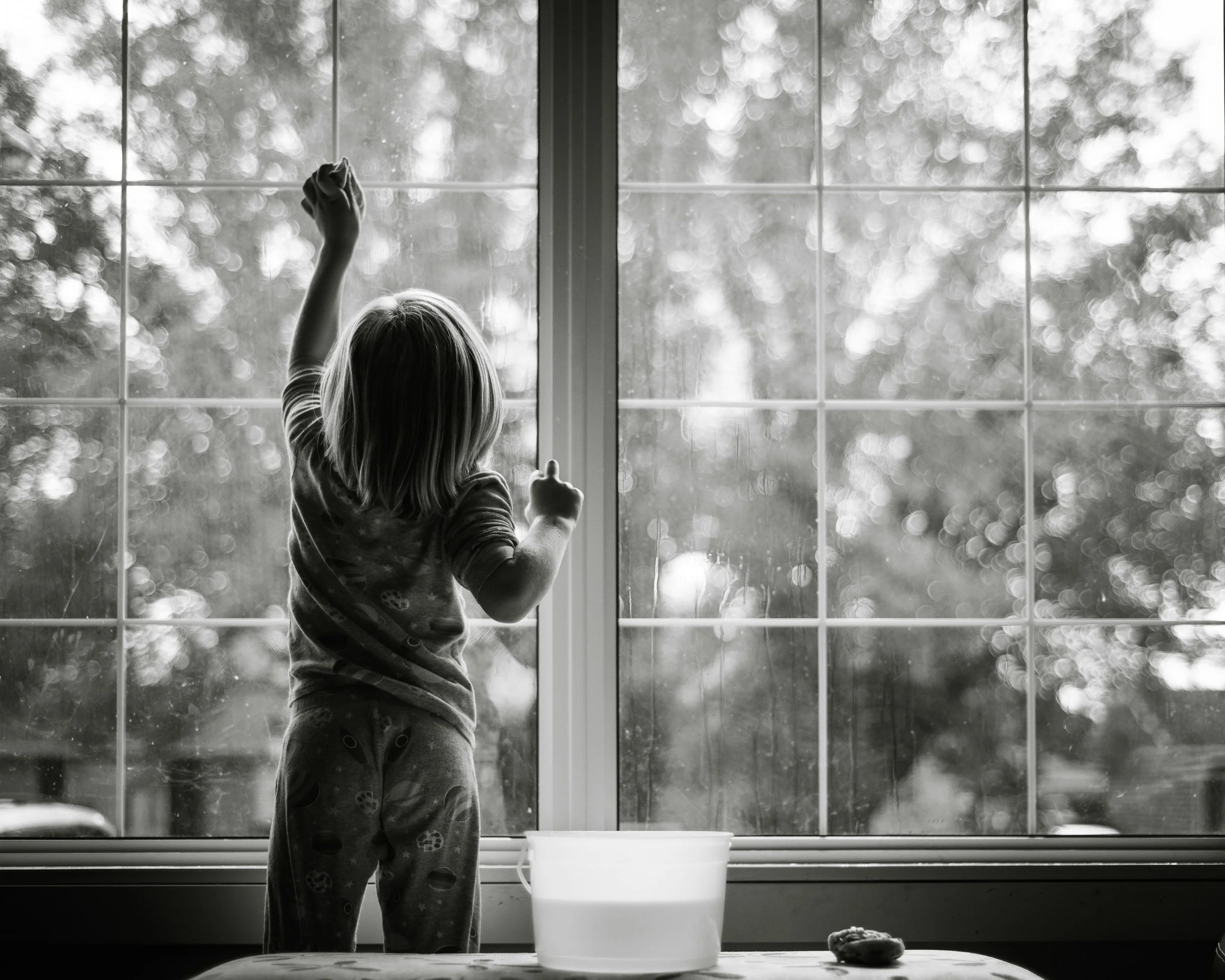A young girl cleaning a large window with a cloth, viewed from inside a home, with a plastic bowl and a small object on a table in the foreground.