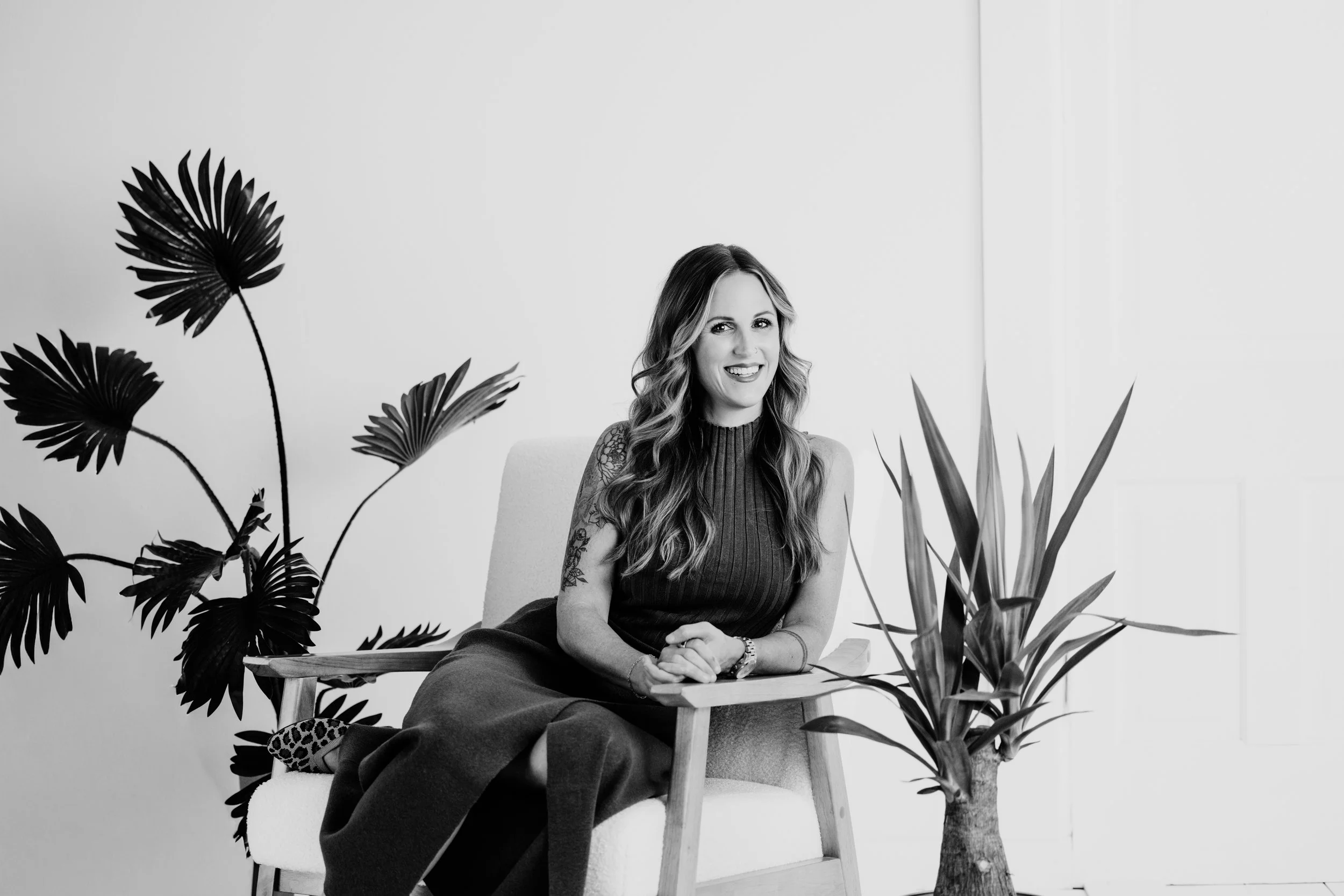 A woman with long wavy hair sitting on a modern chair amidst large tropical plants, smiling and looking at the camera in a minimalist indoor setting.