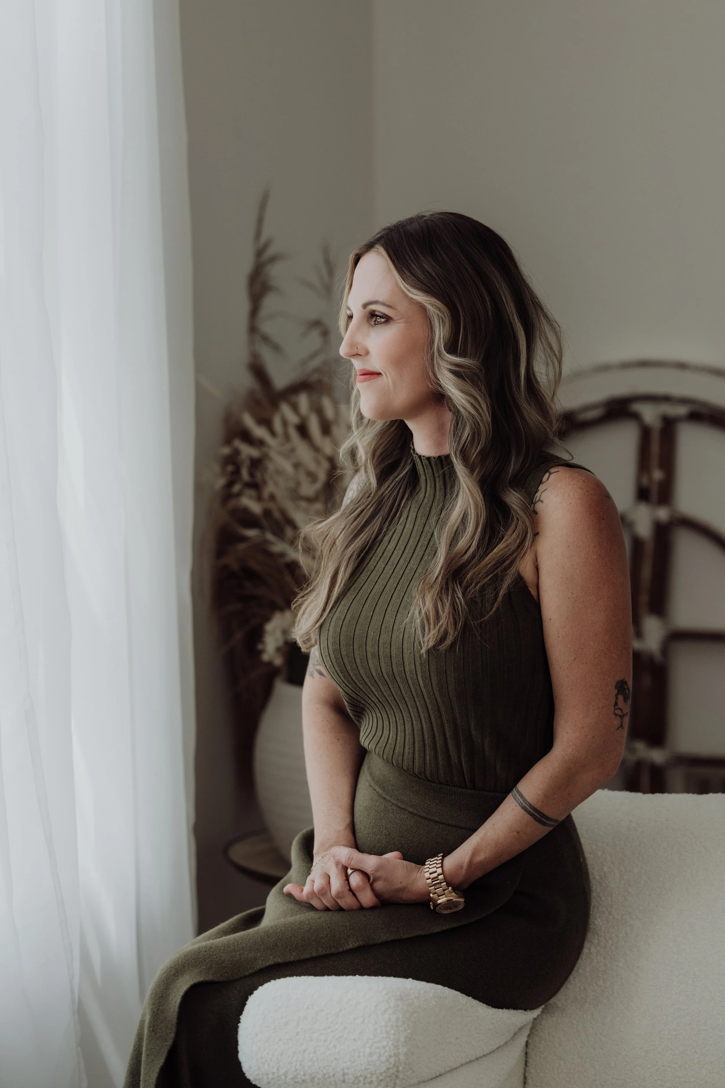 A woman with wavy, shoulder-length hair sitting on a white chair near a window with white curtains, wearing a sleeveless olive green top and matching skirt, with tattoos on her left arm and a watch on her left wrist, looking out the window.