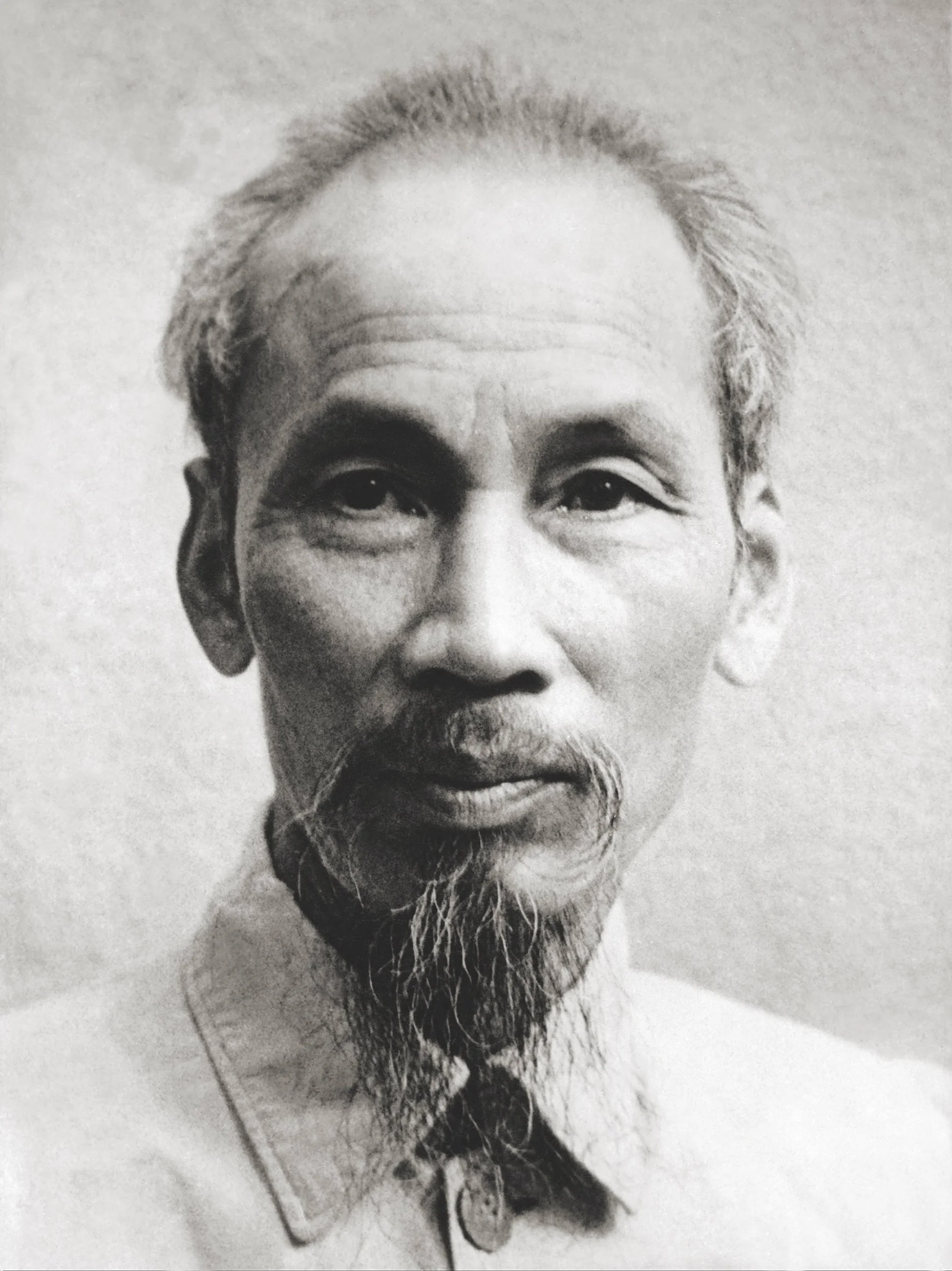 Black and white portrait of Ho Chi Minh, an elderly man with a long beard, high forehead, and thinning hair, wearing traditional clothing with a collar.
