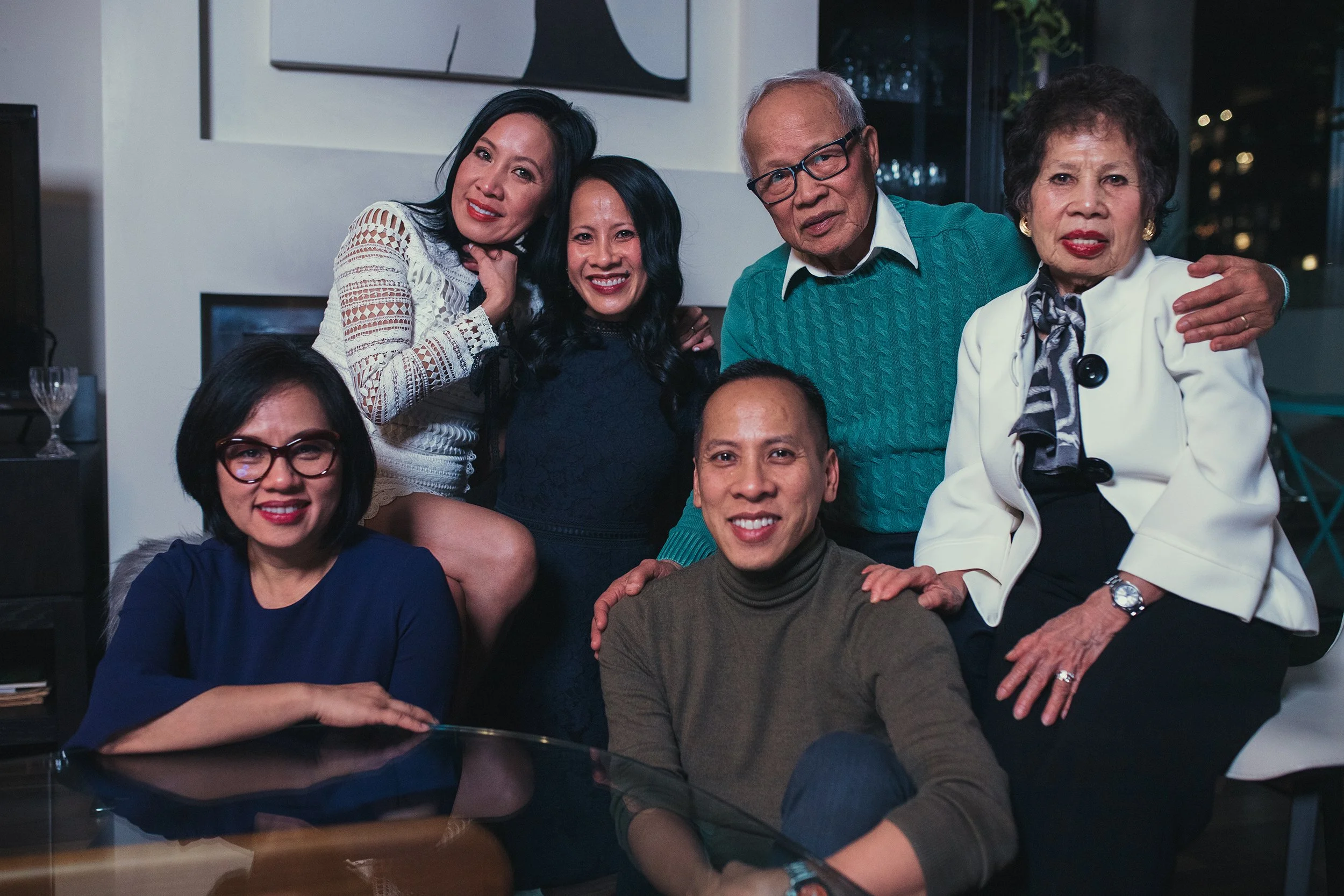 A group of seven family members posing together inside a modern living room, some sitting and some standing, with big smiles, in an evening setting.