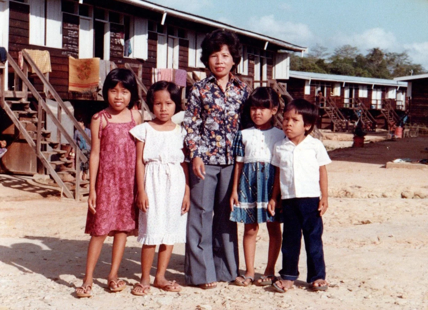 A mother and her four children standing outside in front of wooden houses with stairs and laundry hanging, on a sunny day.