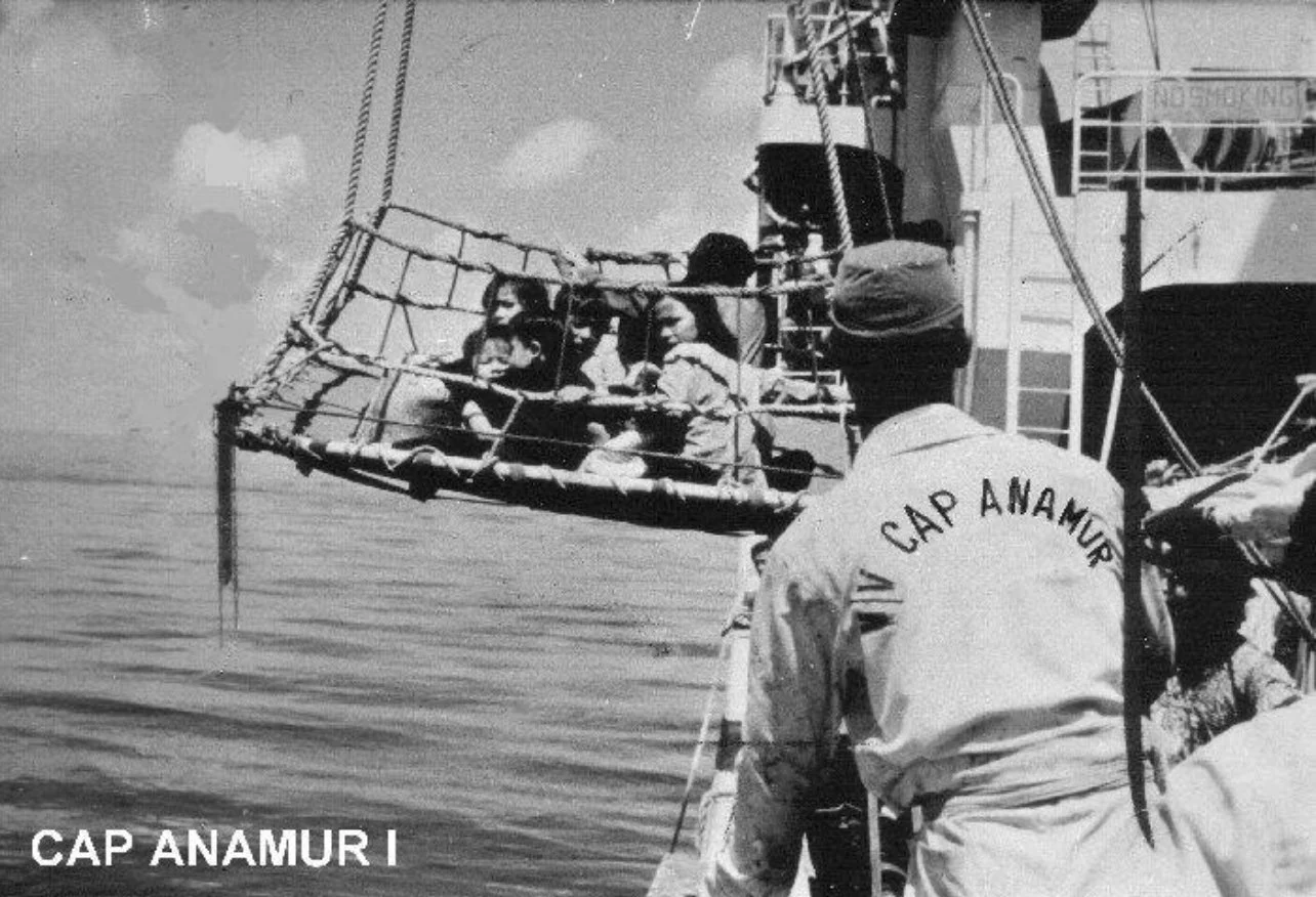 A black and white photo of children sitting in a rescue basket made of wood and rope, being guided by a man in a uniform with 'CAP ANAMUR' written on the back, on calm waters.