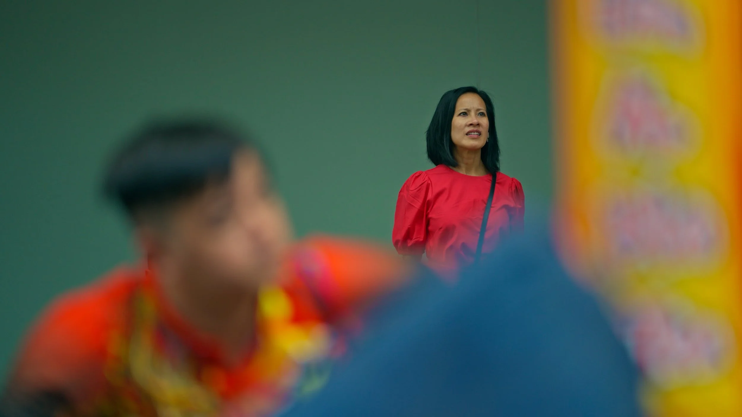 A woman in a red shirt standing behind a blurred person in the foreground near a colorful vertical banner.