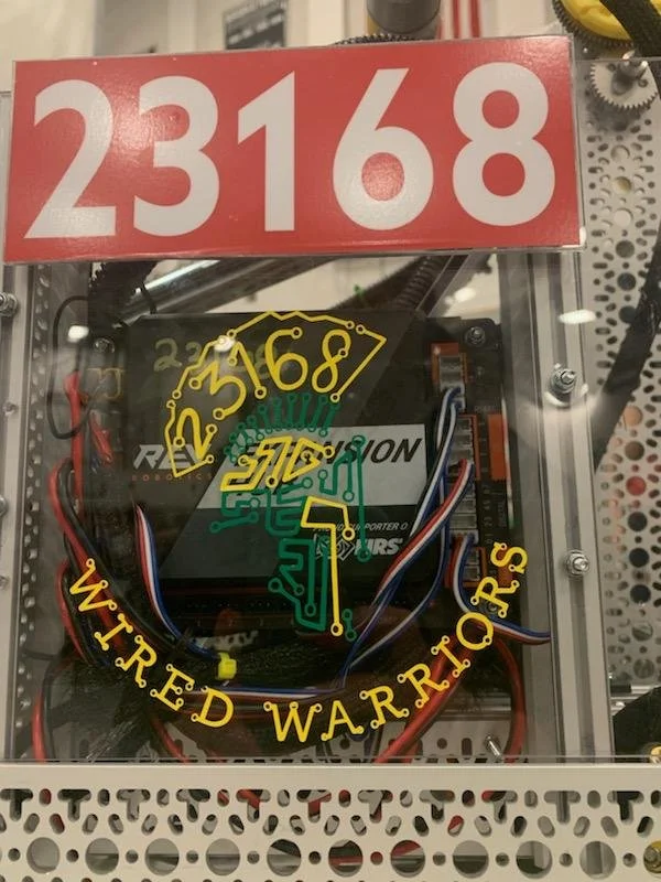 A robotic device with circuitry and wires inside a transparent box. The box has a red label with white numbers "23168" at the top. The front has a yellow nameplate reading "Wired Warriors" with a circuit design.