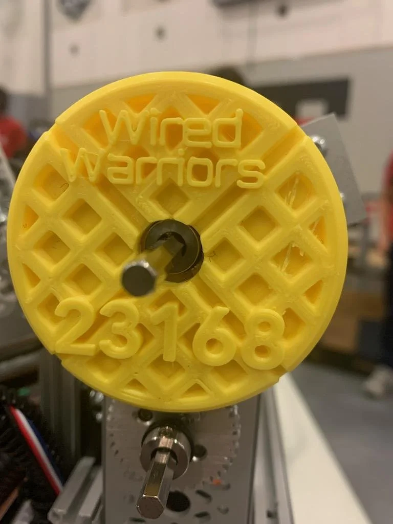A yellow circular object with the words 'Wired Warriors' and numbers 0-9 embedded in it, mounted on a mechanical assembly with a metal lever.