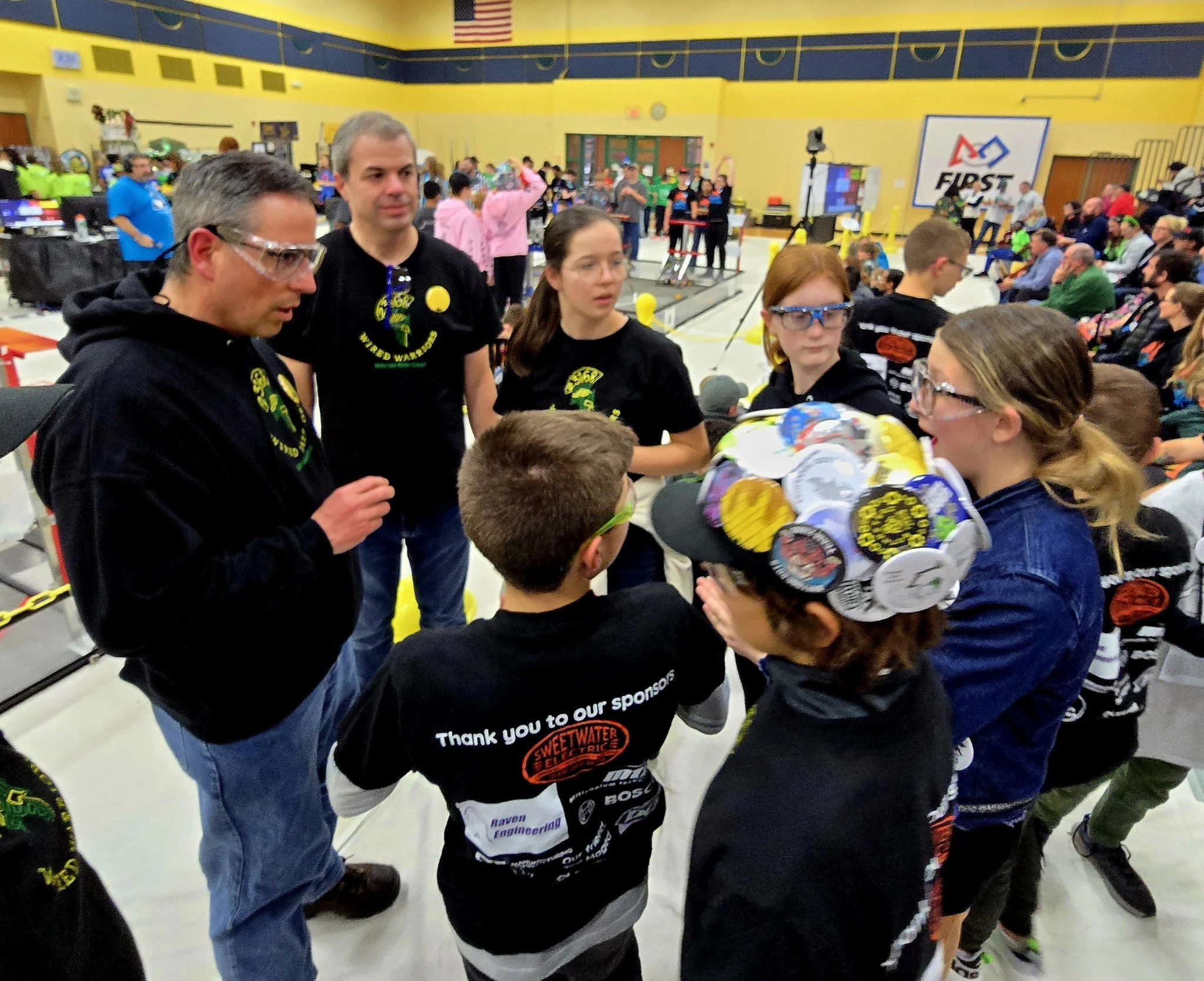 Group of children and adults in a gymnasium during a robotics competition, with some children wearing robot-themed hats, and others wearing shirts with robot logos, seated in the audience and participating in discussions.