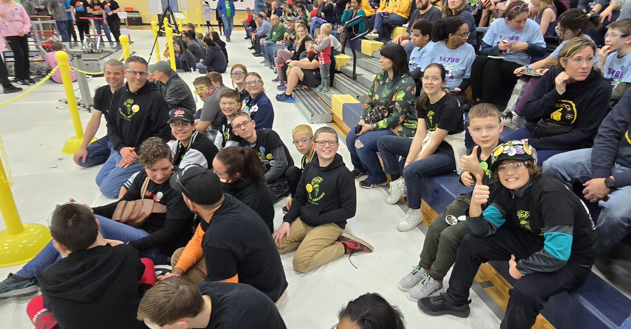 Group of children and adults sitting in bleachers at a school event, some wearing black hoodies with a green and yellow logo, in an indoor gymnasium.