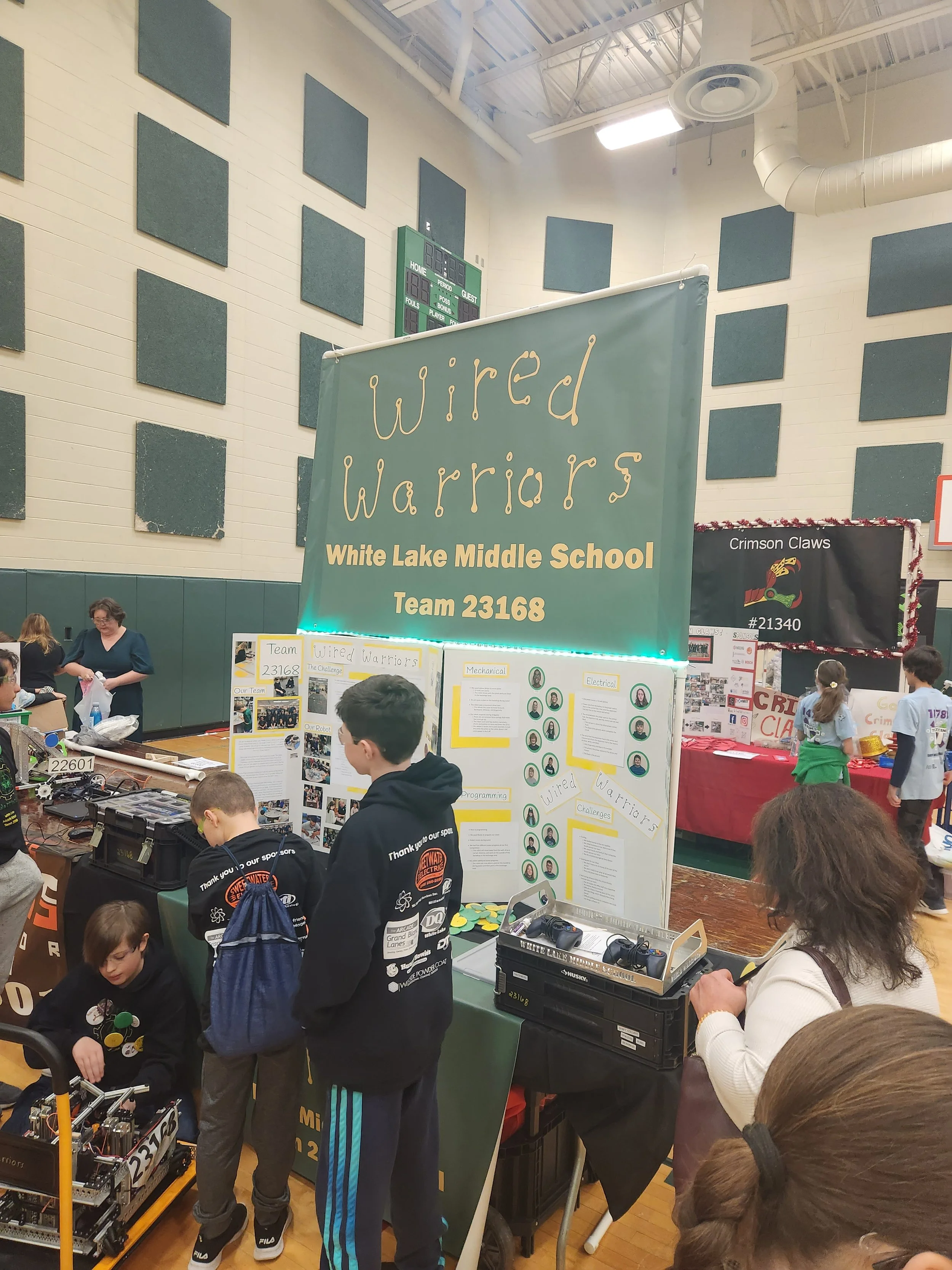 Students and teachers at a school science fair exhibit. A large sign reads 'Wired Warriors' from White Lake Middle School, Team 23168.