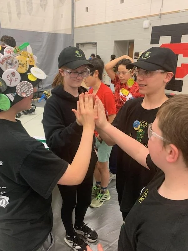 Three kids, two girls and one boy, are playing rock-paper-scissors with each other in a gymnasium. The girl on the left has a cap with colorful buttons, the girl in the middle is wearing glasses and a black hoodie, and the boy on the right is wearing