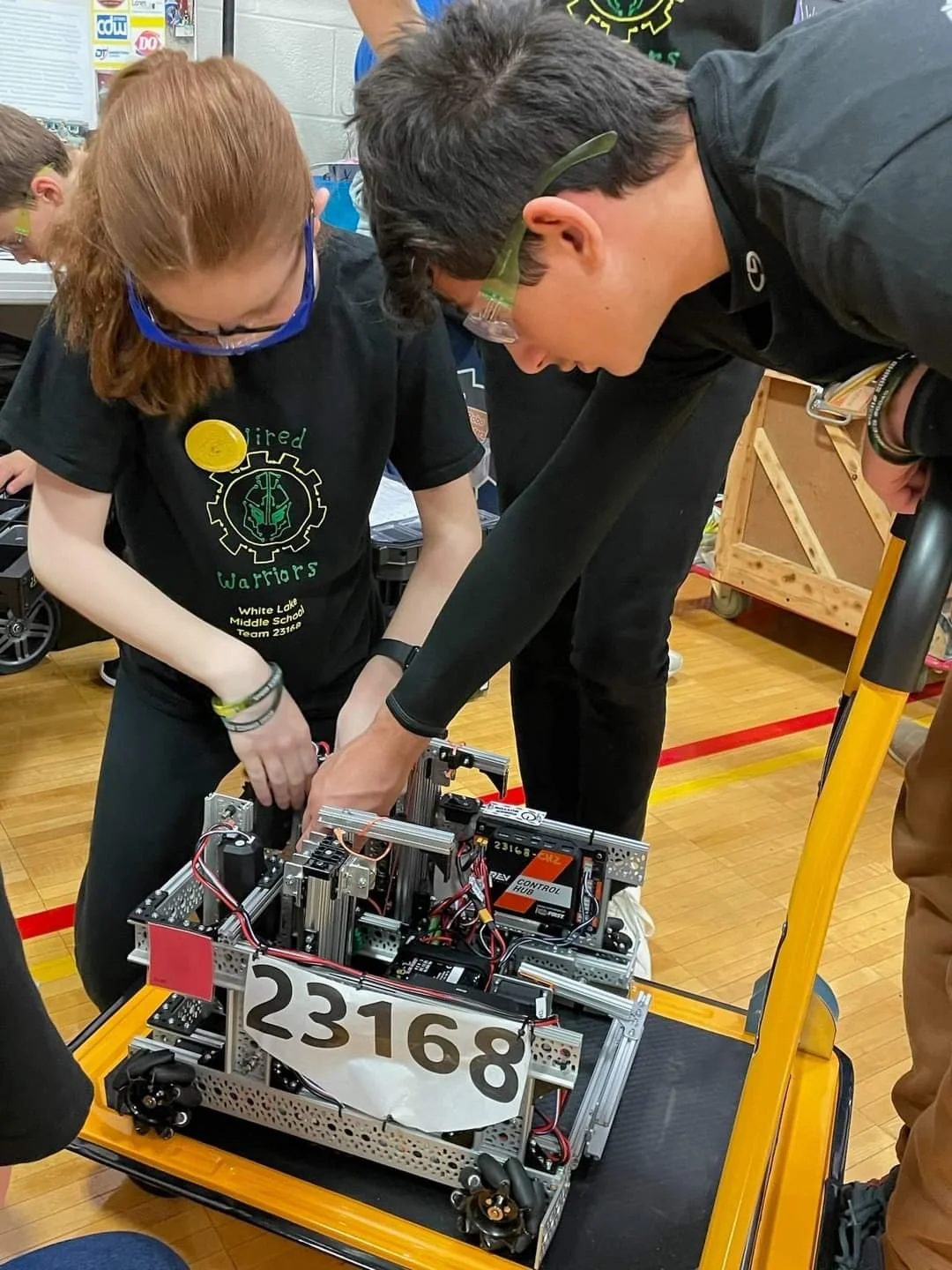 Two students working together on a robotics project in a school gymnasium, with a robot labeled '23168' on a yellow cart.