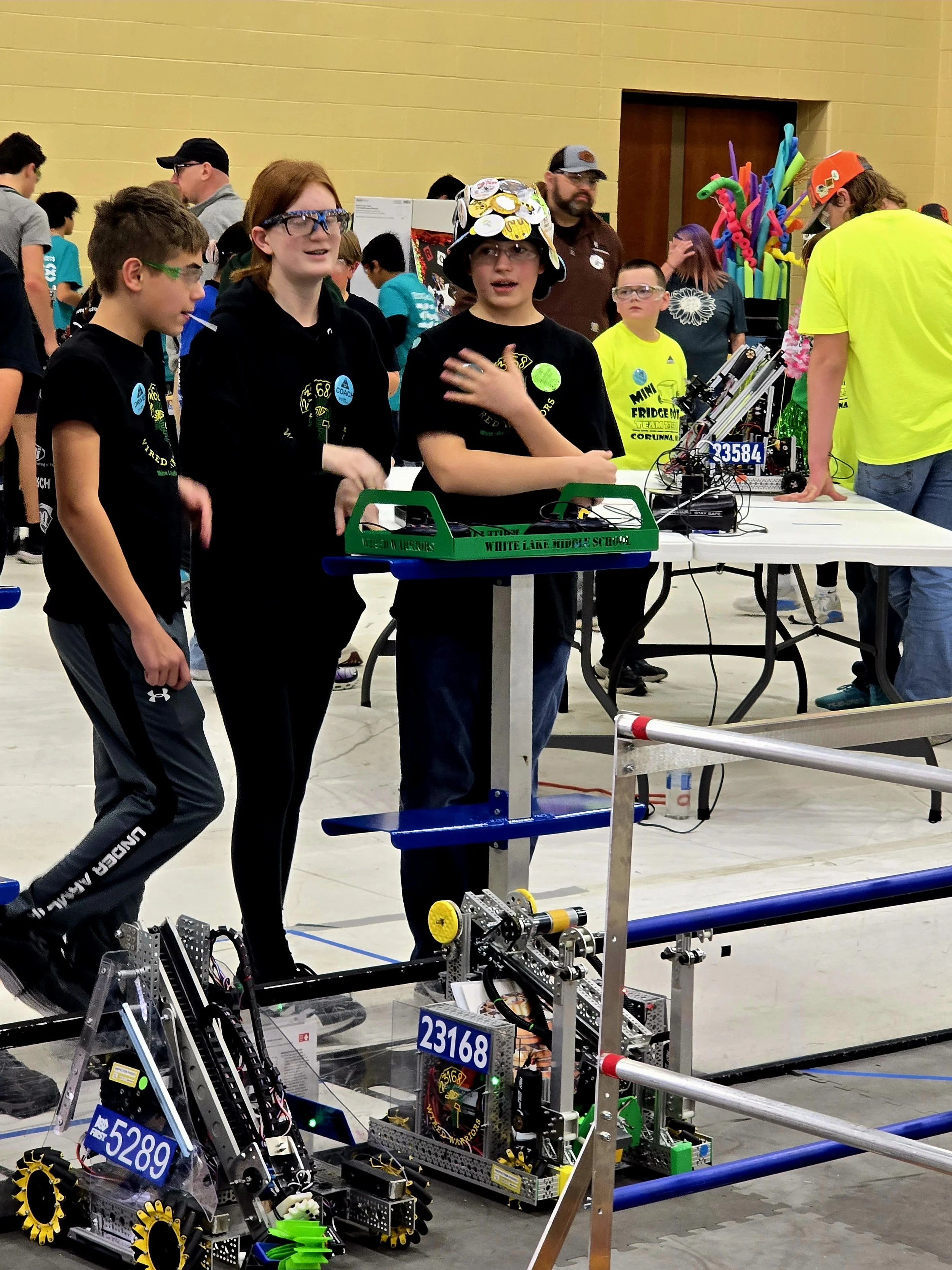Young students participating in a robotics competition, standing next to their robots with a crowd in the background.