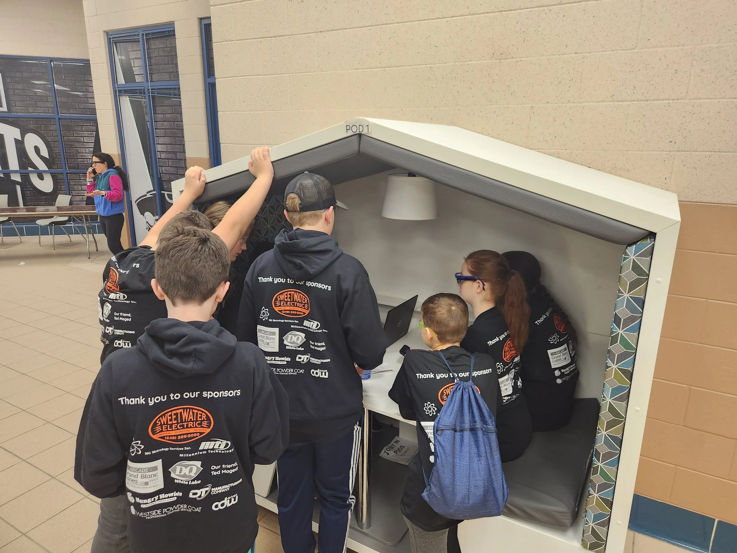 Group of children gathered around a small interactive booth with a woman, inside a school hallway.