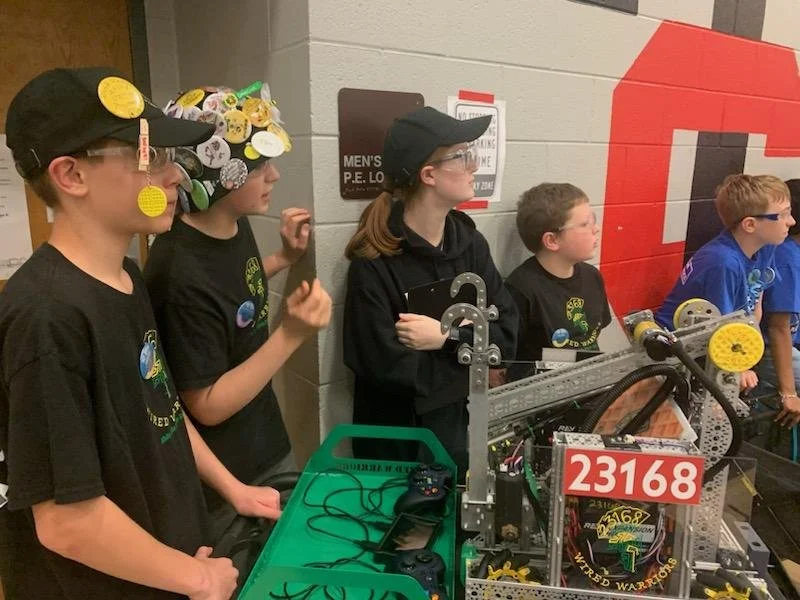Group of children in black and blue shirts with goggles and hats, standing behind a workbench with robotics equipment, inside a school or community center gymnasium.