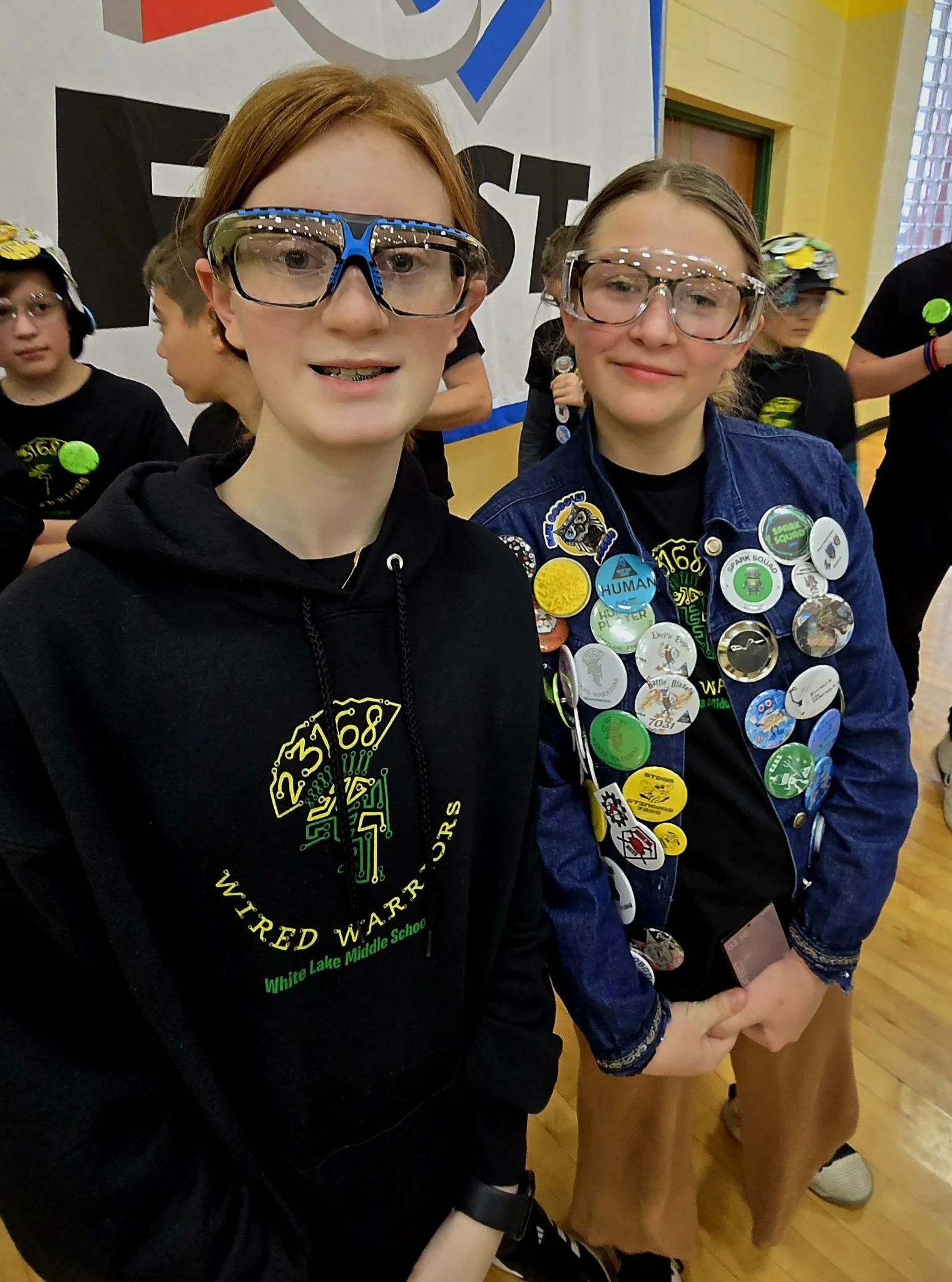 Two girls at a school event, both wearing glasses and decorated with numerous badges. The girl on the left has red hair and is wearing a black hoodie, while the girl on the right has brown hair, a denim jacket, and tan pants. They are standing indoor