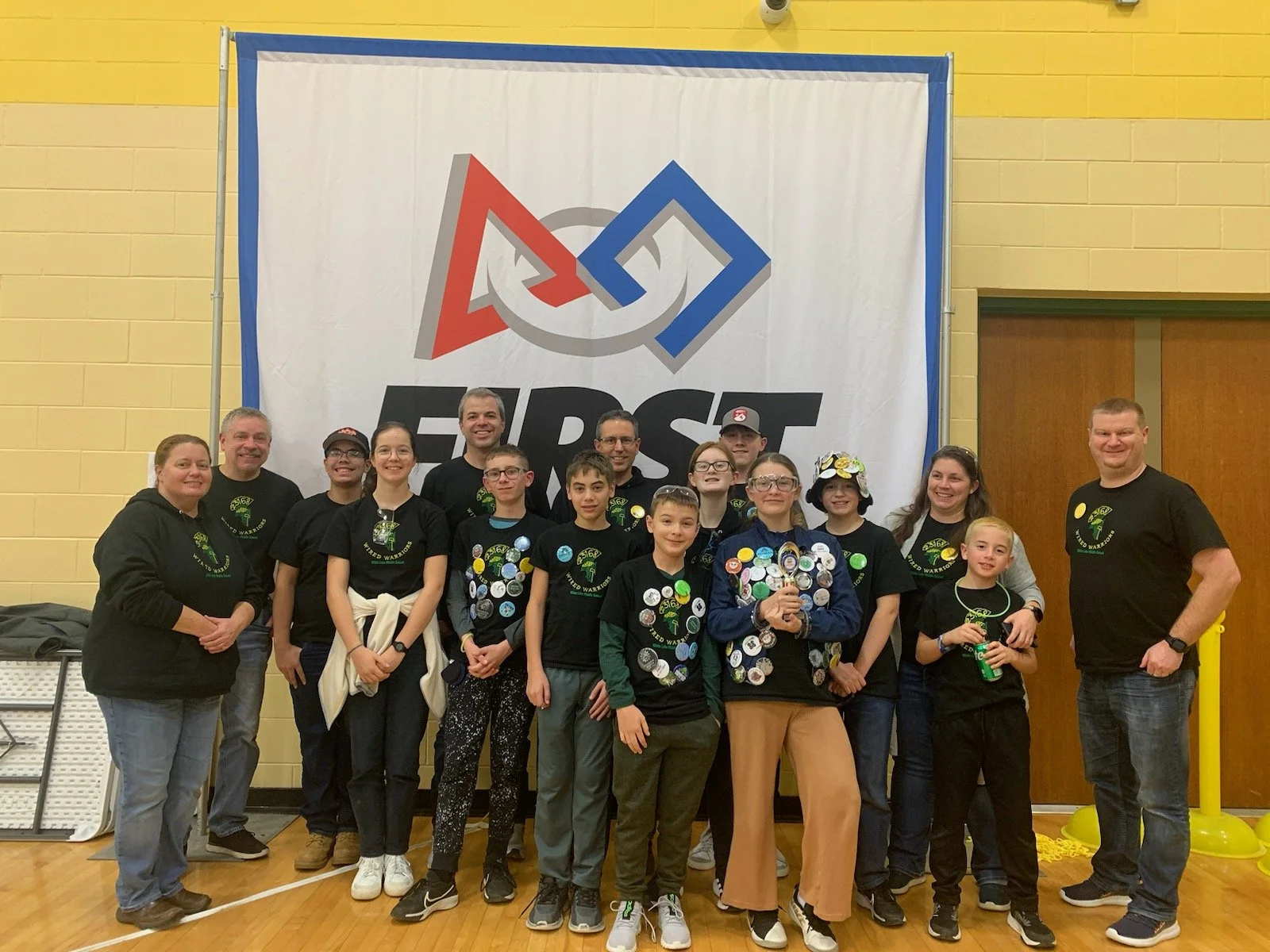 Group of young people and adults smiling in front of a large banner with a logo and the word "FIRST" at a robotics competition.