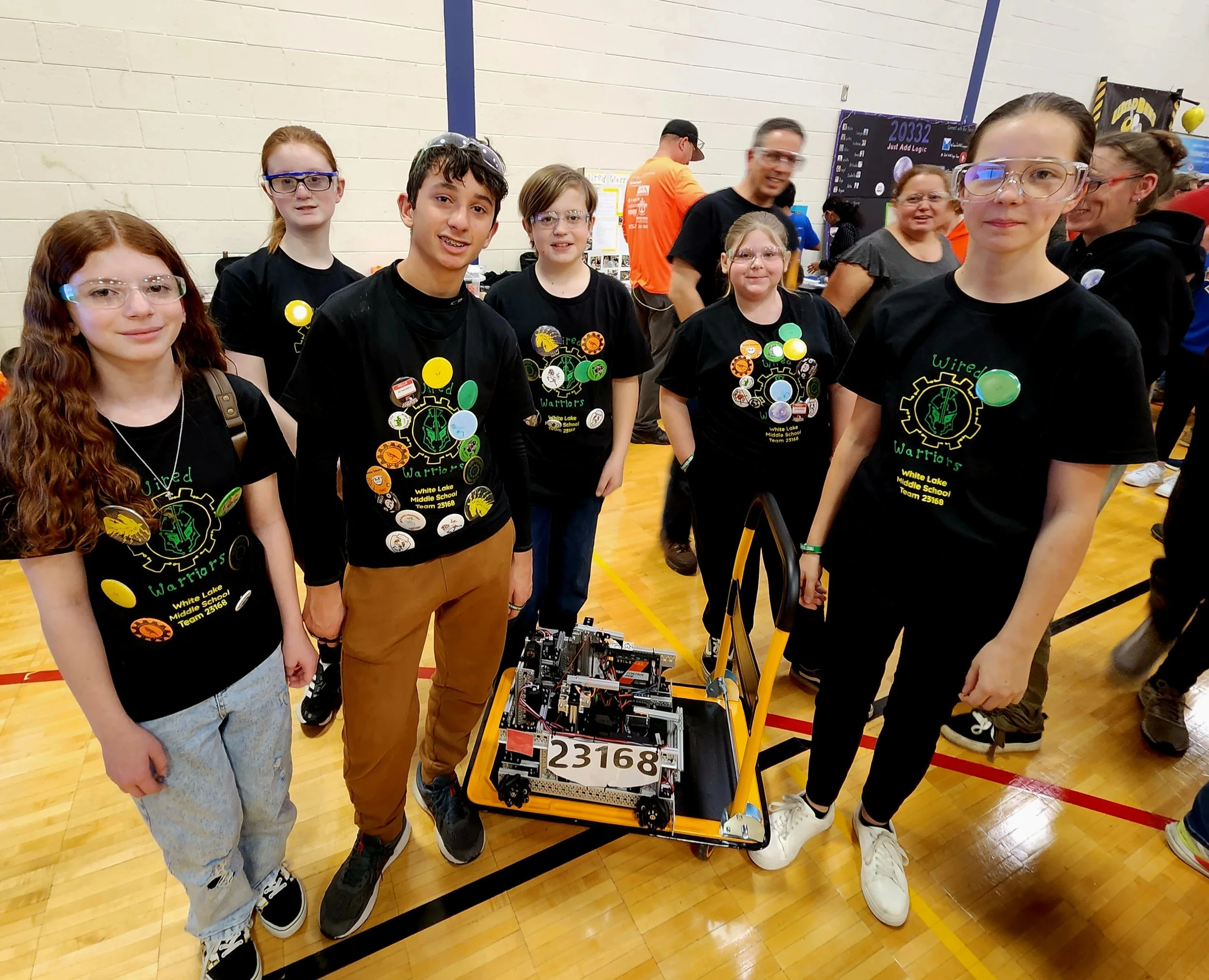 Group of middle school students wearing matching black T-shirts with green and yellow robot logo, posing in a gymnasium, standing behind a robot with number 23168 for a robotics competition.
