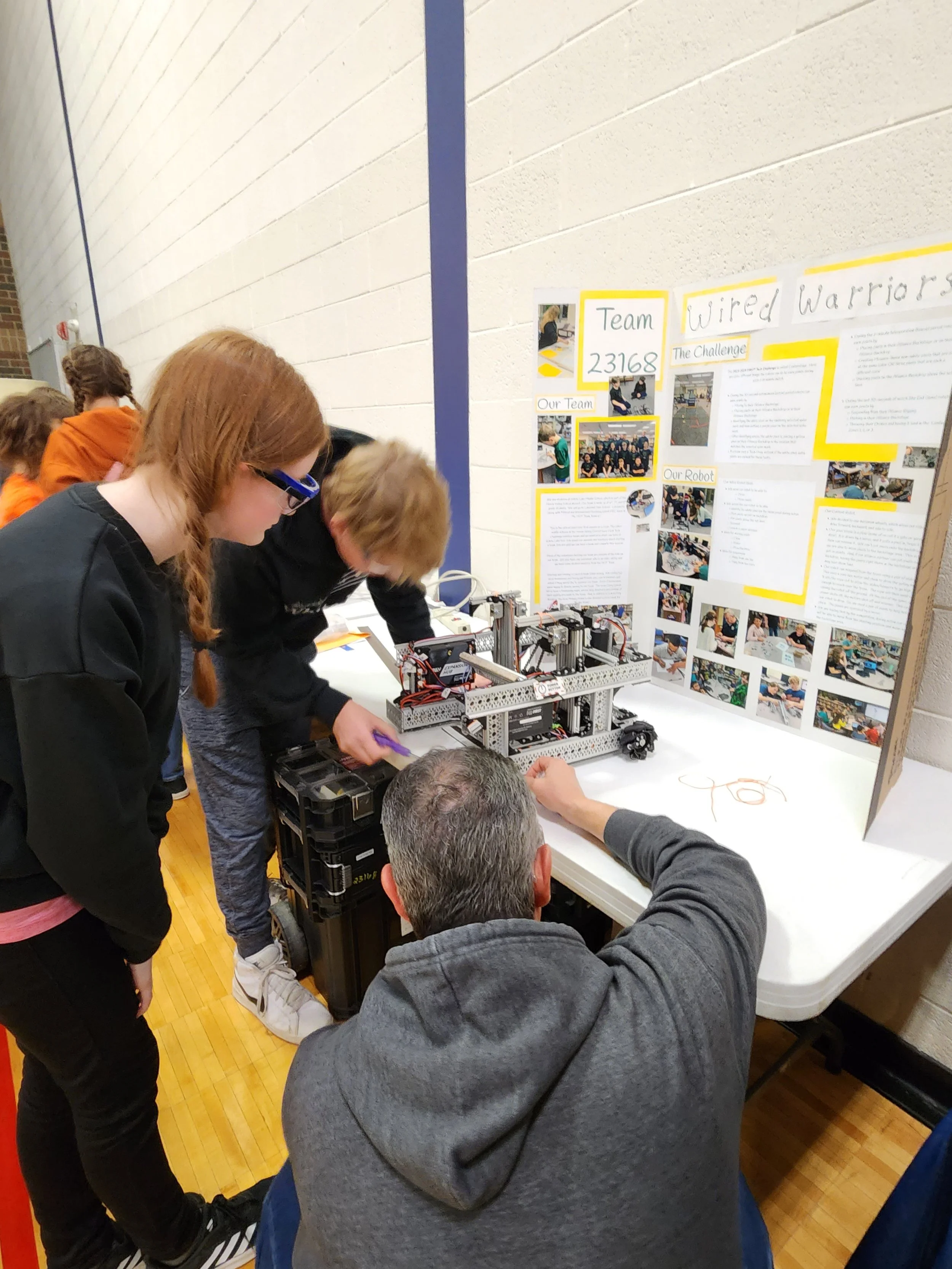 Students and a teacher working on a robotics project at a school event, with a display board titled 'Wired Warriors' and team information in the background.