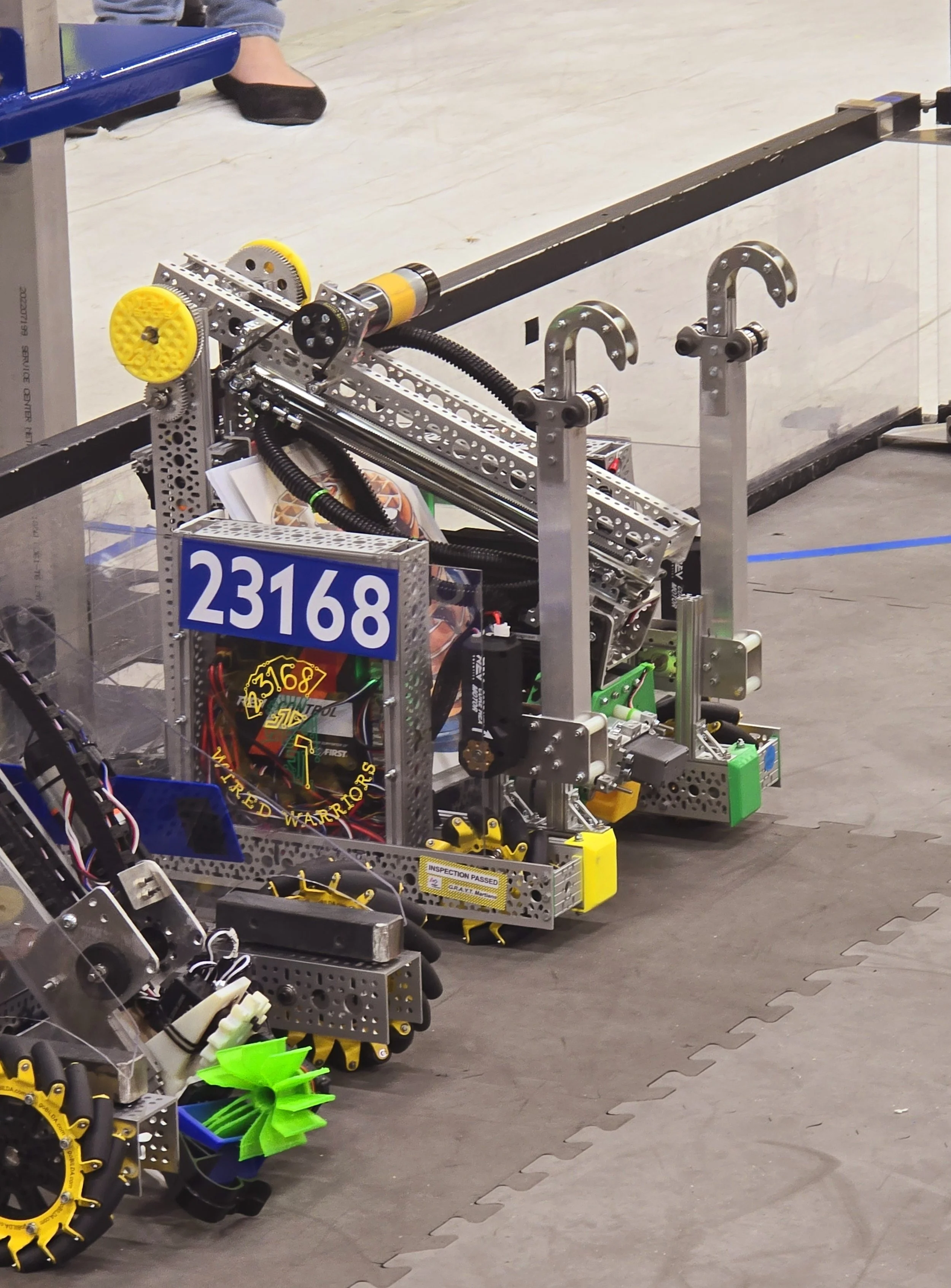 A robot built with metal parts, wires, and gears is on a gray mat. It has a blue number 23168 on its side and a yellow gear with the words "Wired Warriors" written on it. The robot appears to be part of a robotics competition or event.